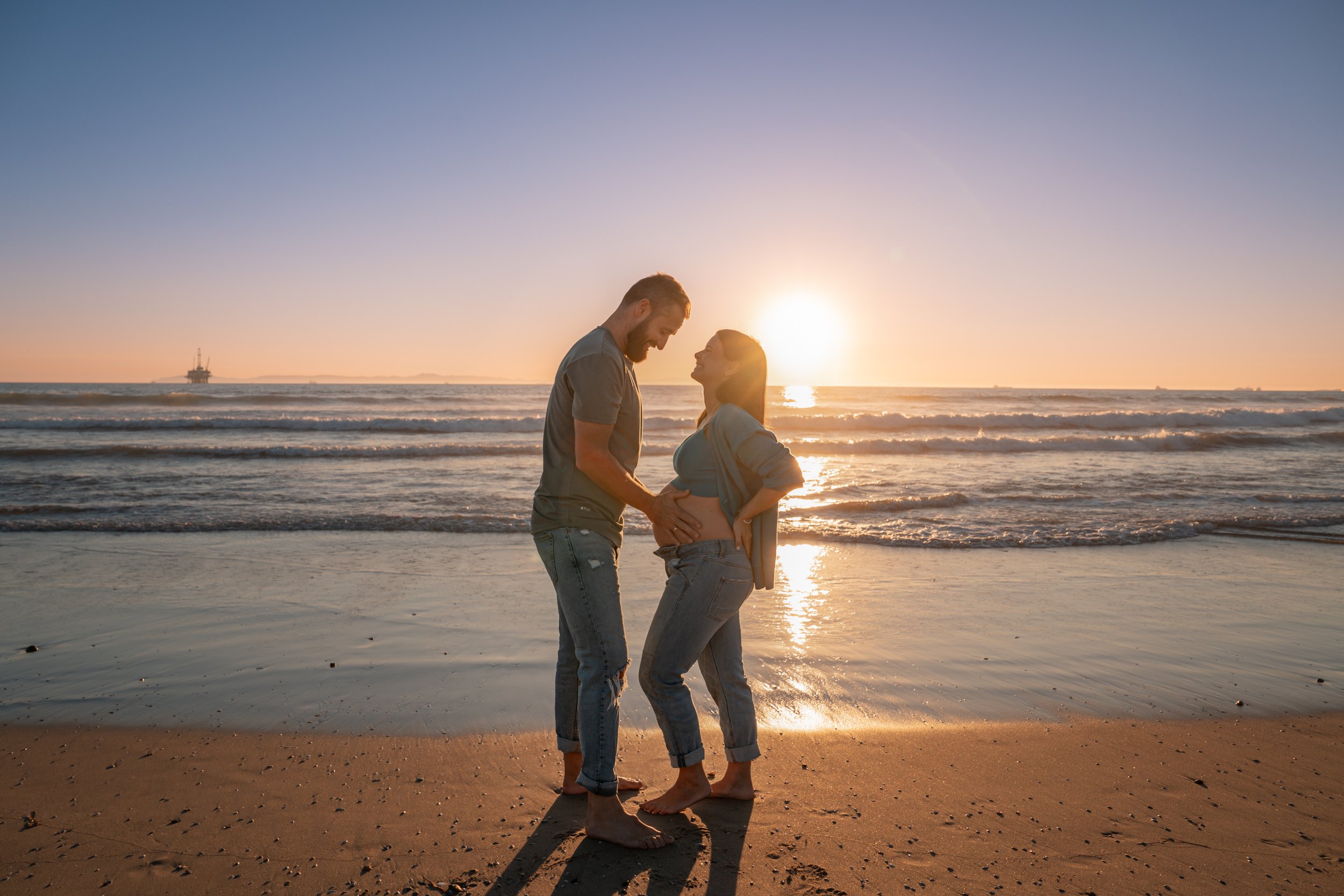 A couple on the beach at sunset, with the man touching the woman's pregnant belly, both smiling at each other.