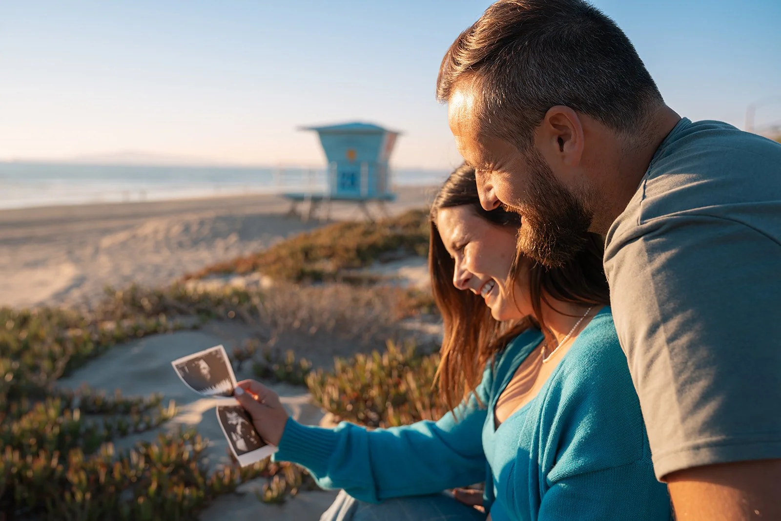 A couple sitting on the beach, smiling and looking at ultrasound images, with a lifeguard tower and sandy shoreline in the background during sunset.