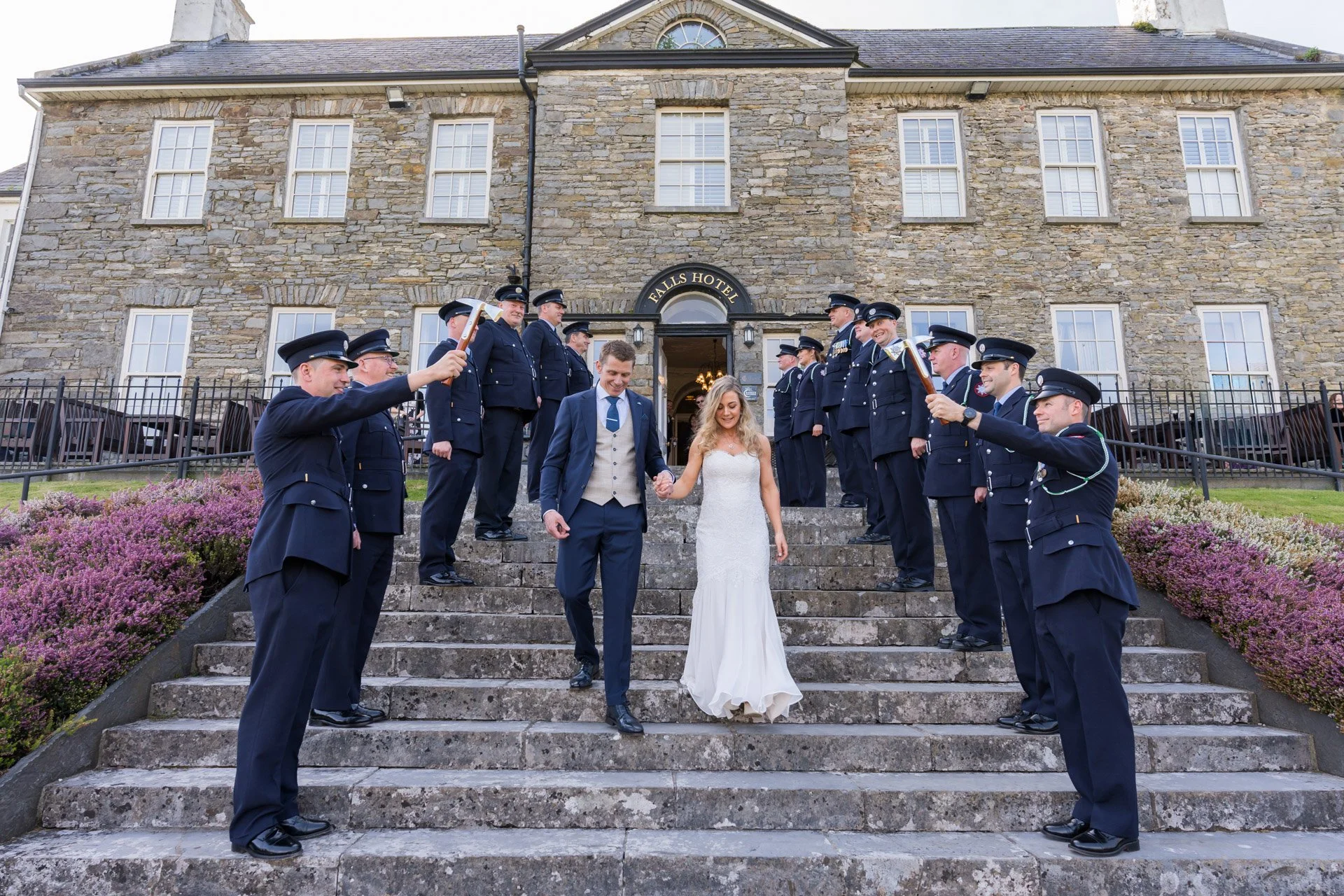 Bride and groom descending stone stairs outside a historic hotel, surrounded by uniformed police officers forming a celebratory arch, with pink flowering bushes on either side.