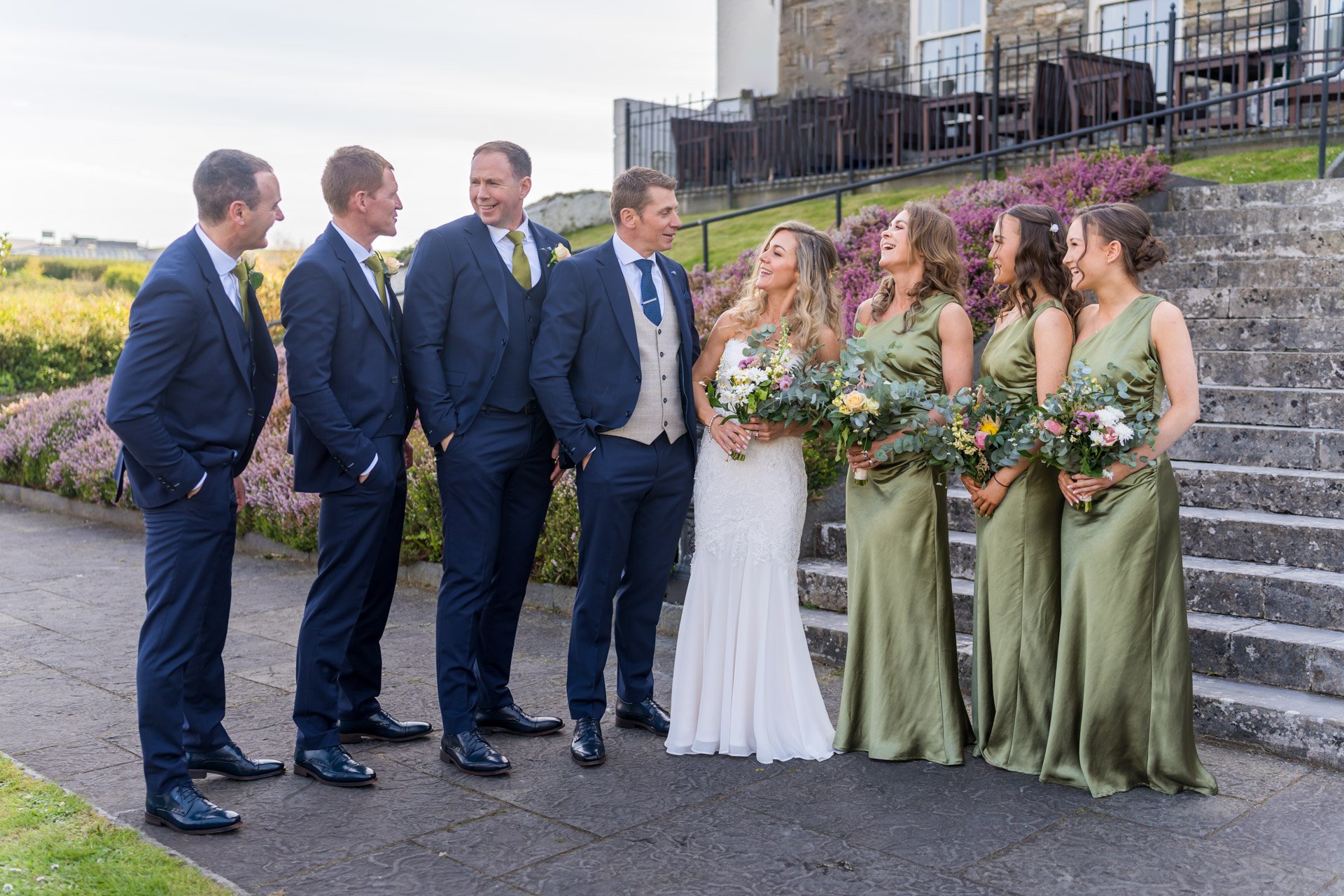 Group of people at a wedding, dressed in formal attire, standing outdoors on stone pavement. The groom and bride are in the center, surrounded by bridesmaids and groomsmen, with a garden and steps in the background.