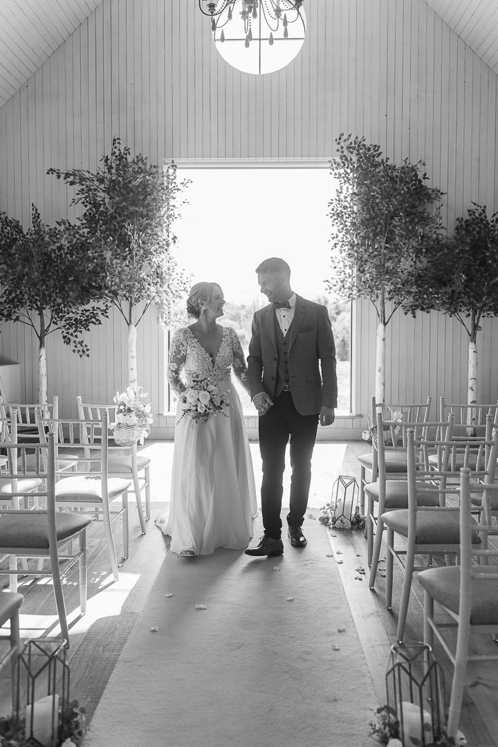 A bride and groom walking down the aisle inside a decorated wedding venue with chairs, trees, and floral arrangements, backlit by a large window.