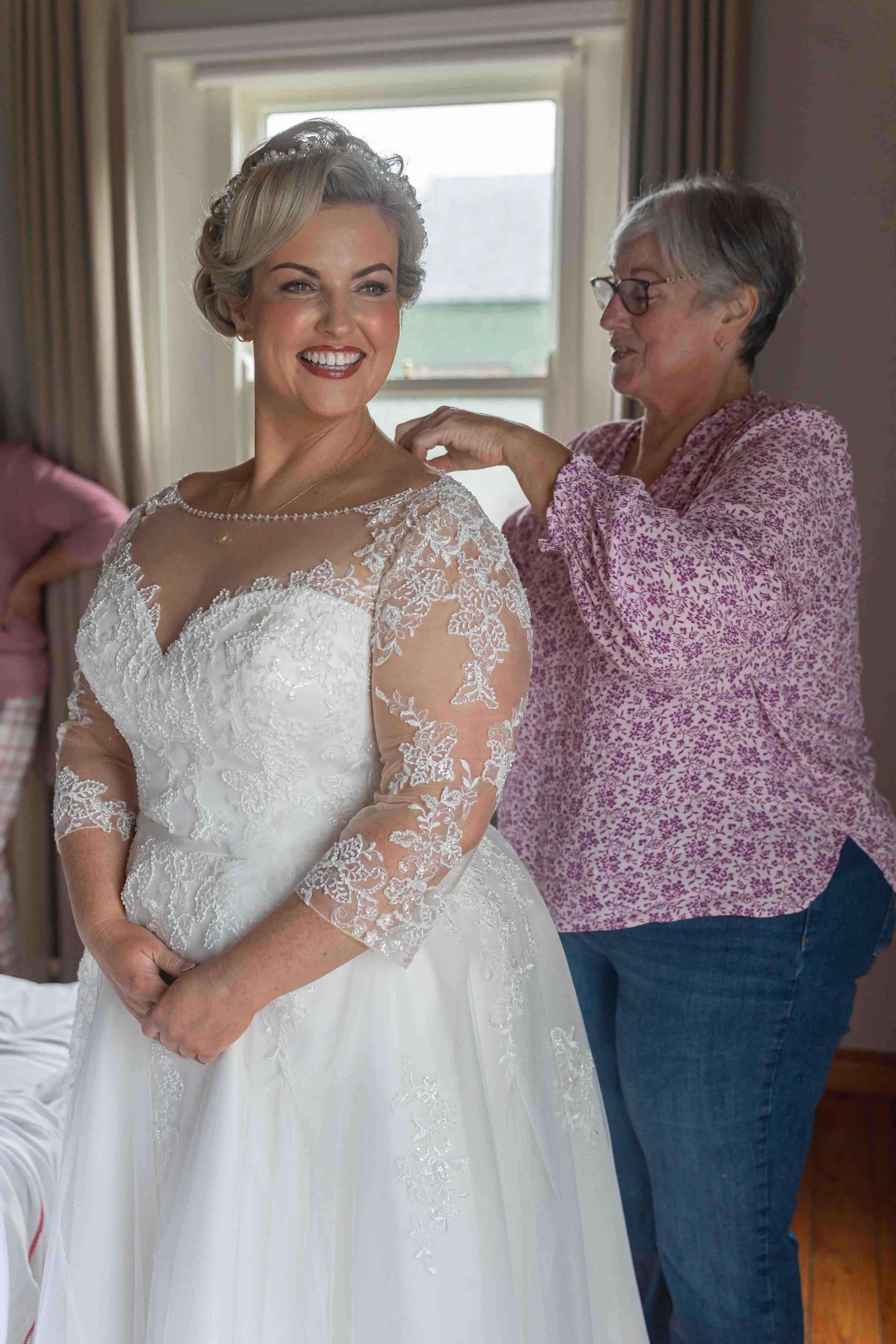 A bride laughing while getting dressed in her wedding gown, assisted by an older woman in a floral blouse, near a window with sunlight.