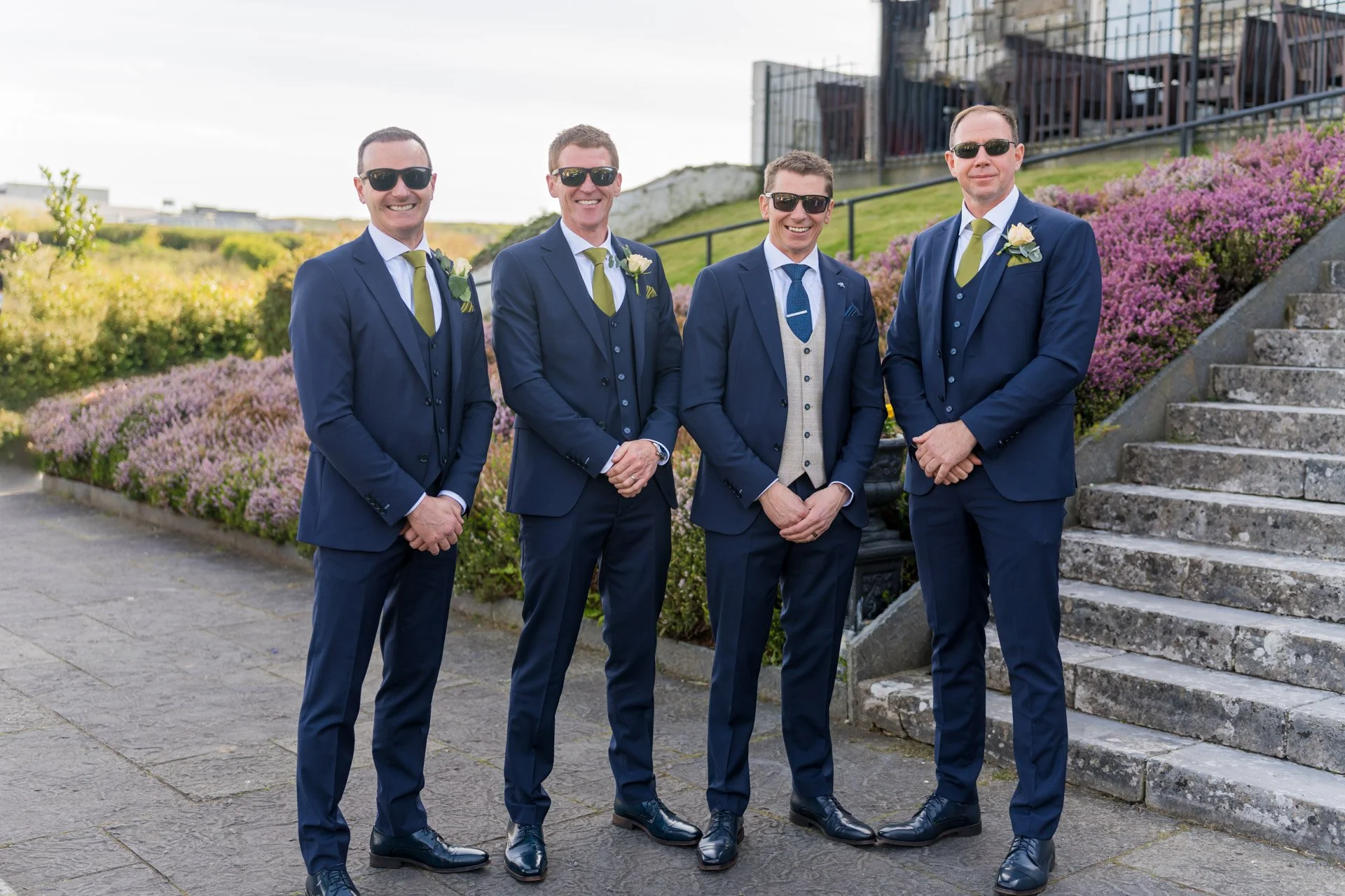 Four men in dark blue suits with white shirts and green ties, wearing sunglasses, standing outdoors near a stone staircase and purple shrubbery, smiling at the camera.