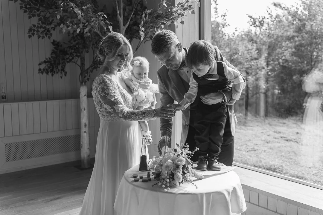 A family celebrating at a wedding, with a woman in a lace wedding dress, a man in a suit, a young boy, and a small girl, all gathered around a table with a flower arrangement, near a large window overlooking an outdoor scene.