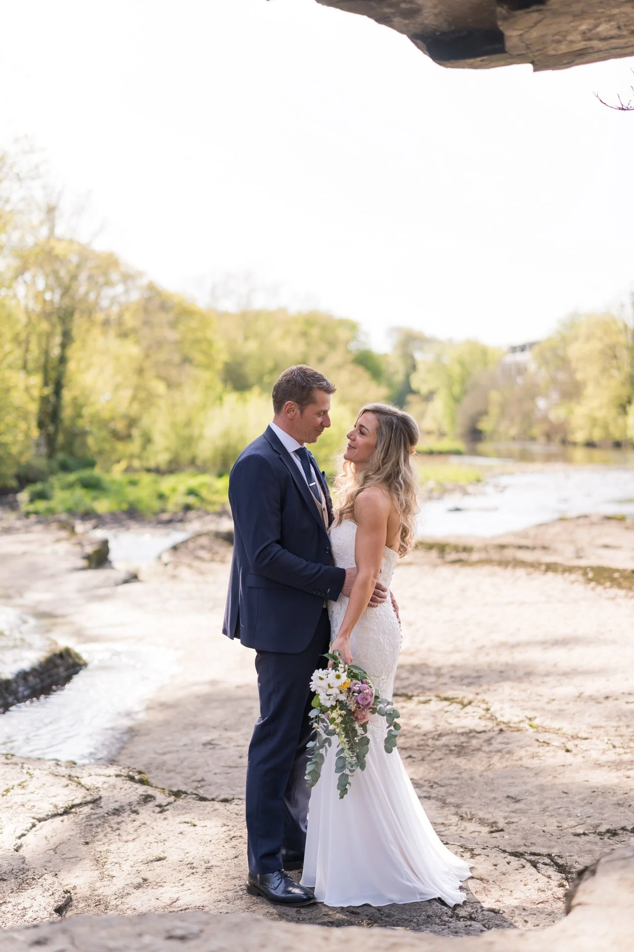 A bride and groom standing close together outdoors by a river, holding hands and gazing into each other's eyes, with trees and a rocky stream in the background.