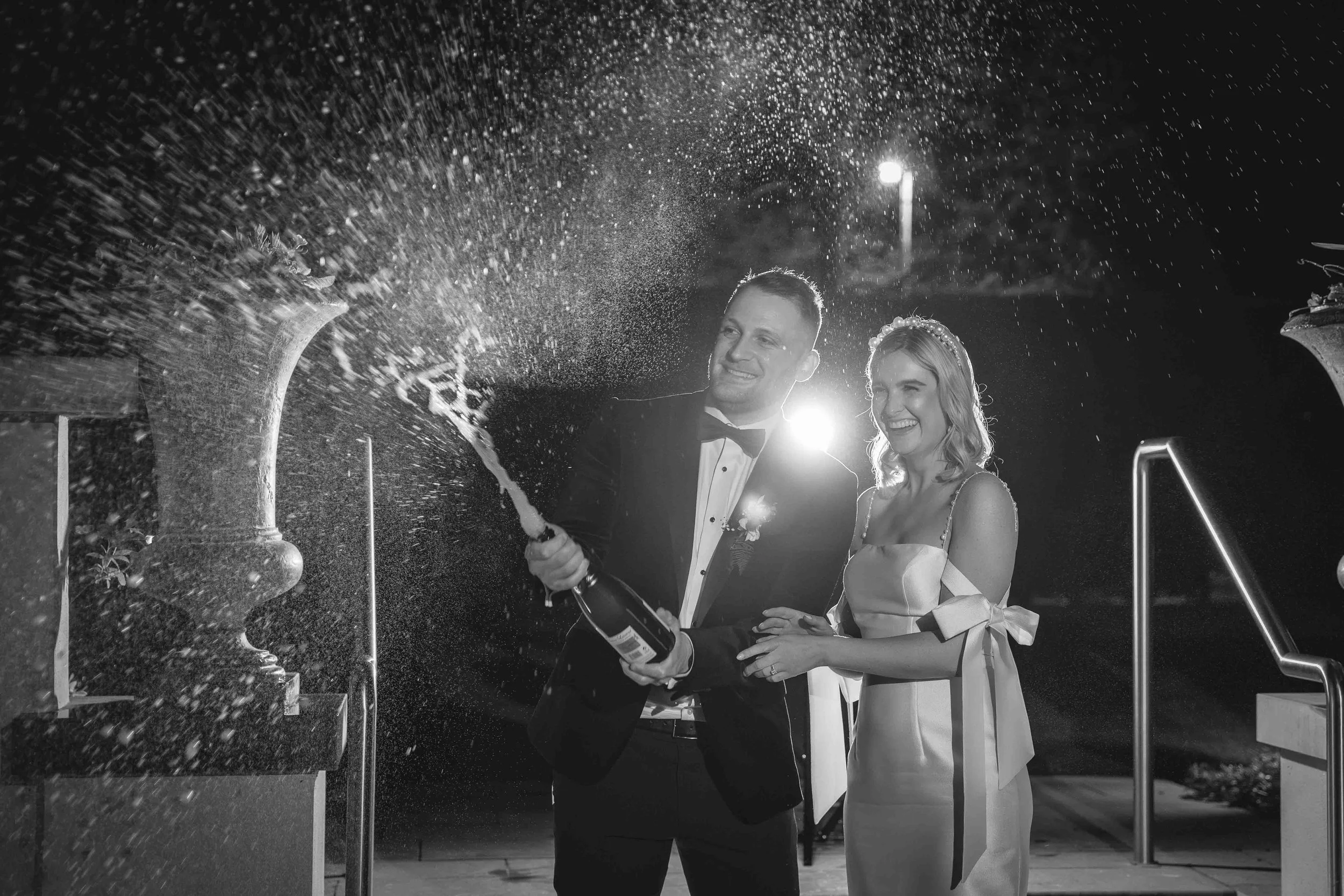 A newlywed couple in formal attire celebrating with champagne, smiling as the groom opens the bottle, causing champagne to spray.