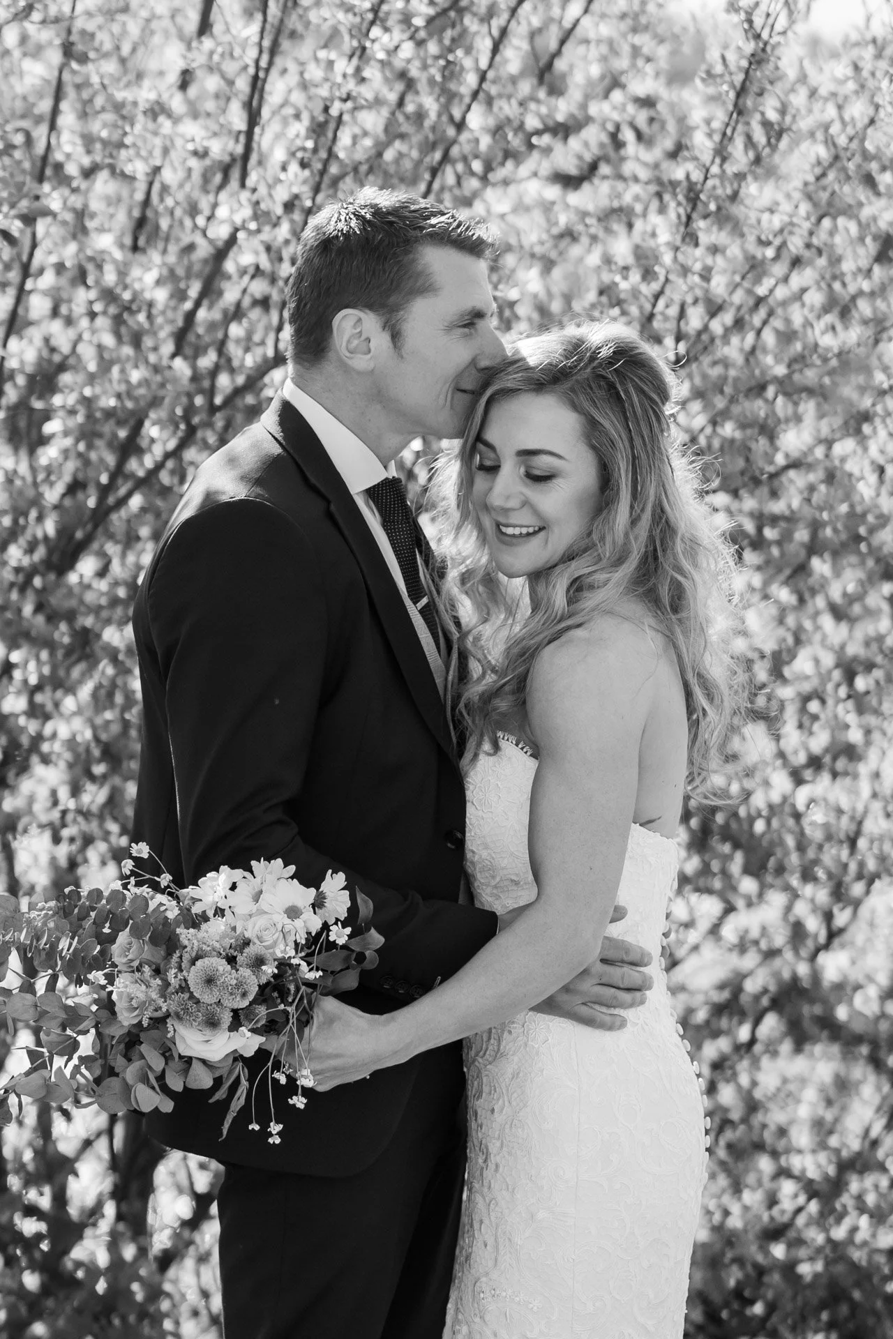 A black and white photo of a bride and groom embracing outdoors. The groom is kissing the bride on her forehead while she smiles, holding a bouquet of flowers.