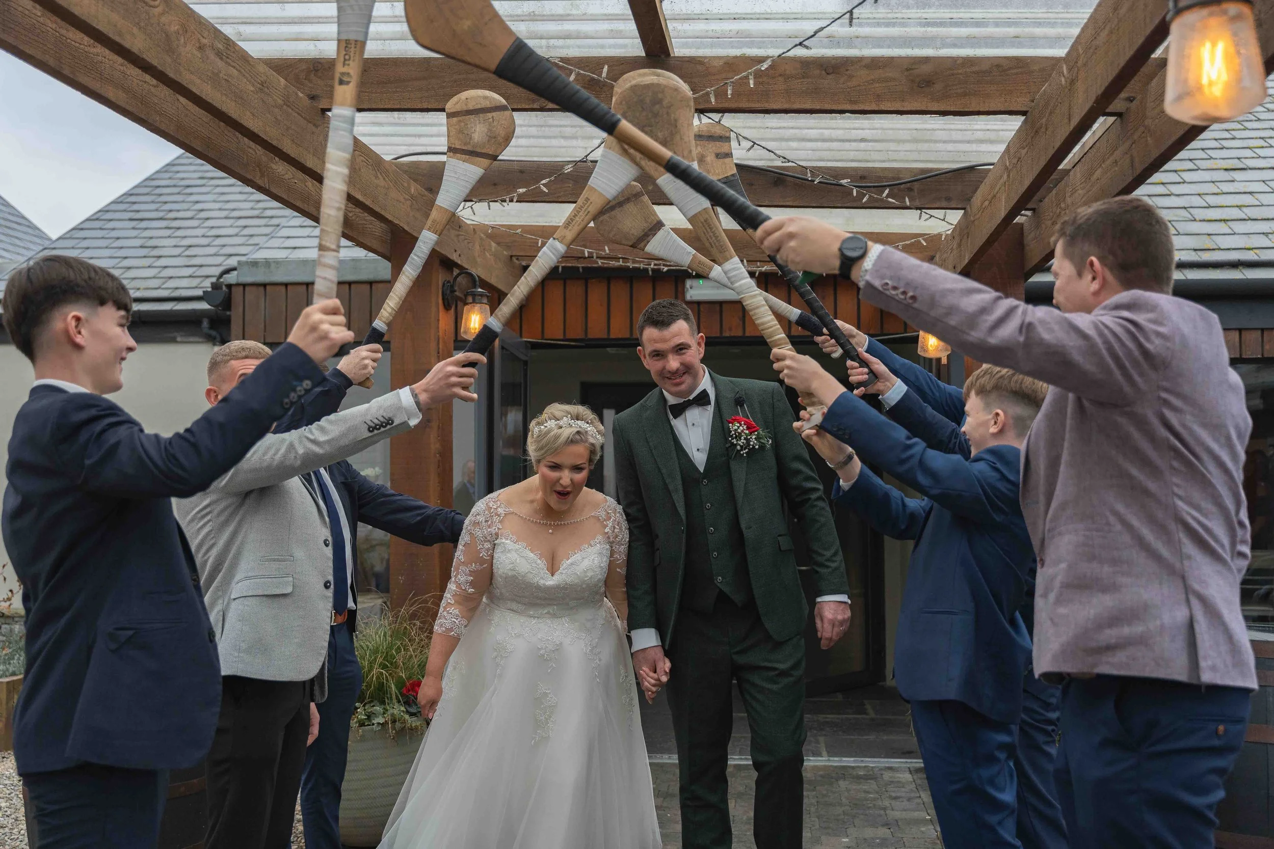 Bride and groom walking together through a tunnel of people holding baseball bats overhead at a wedding reception.