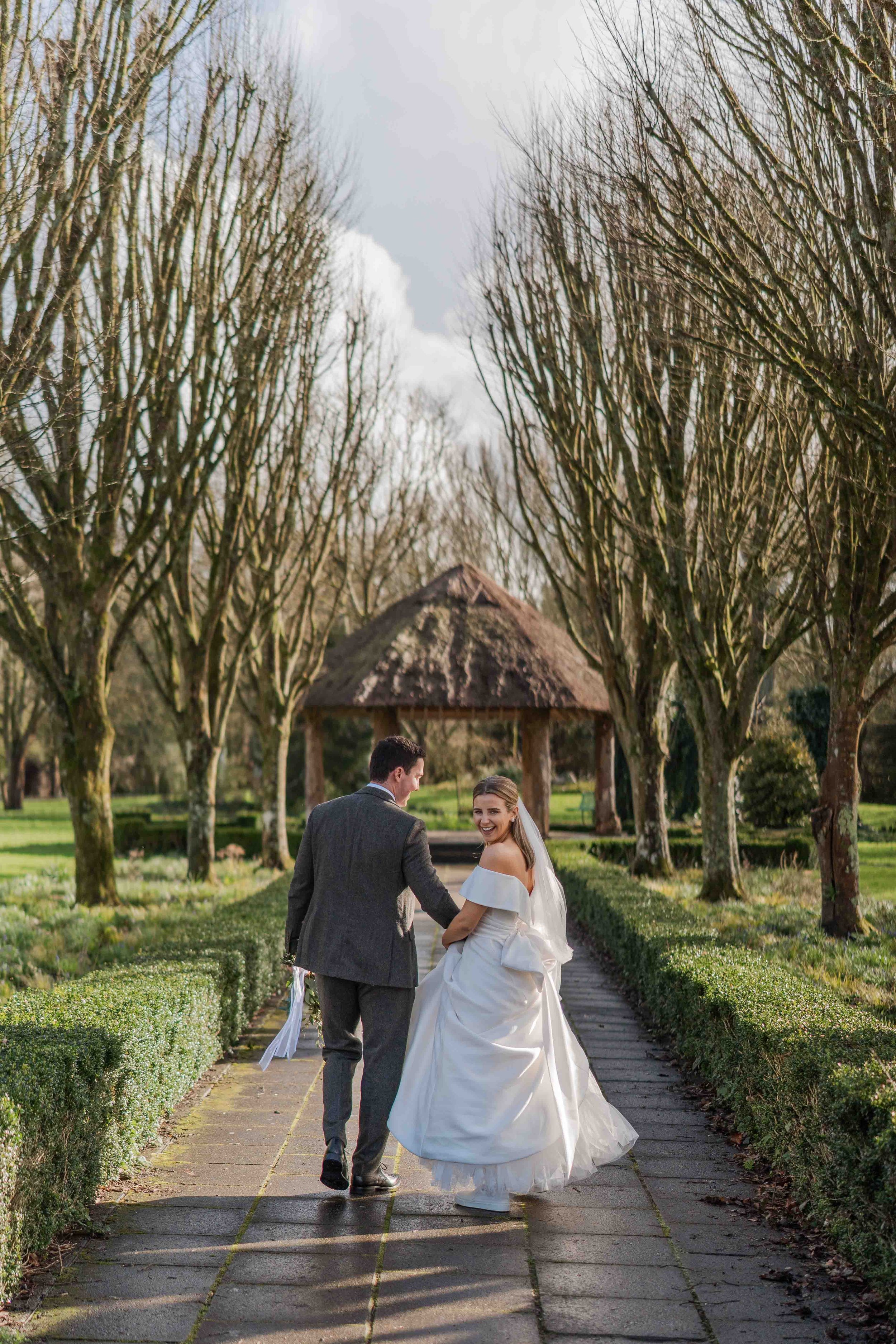 A bride and groom walking hand in hand on a garden path, smiling, with trees and a thatched gazebo in the background on a bright day.