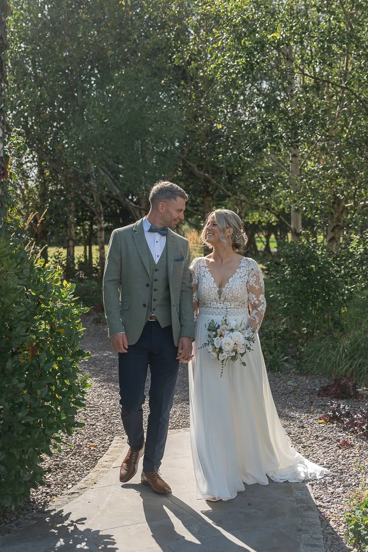 Bride and groom holding hands walking on a garden pathway, smiling at each other on their wedding day, surrounded by greenery and sunlight.