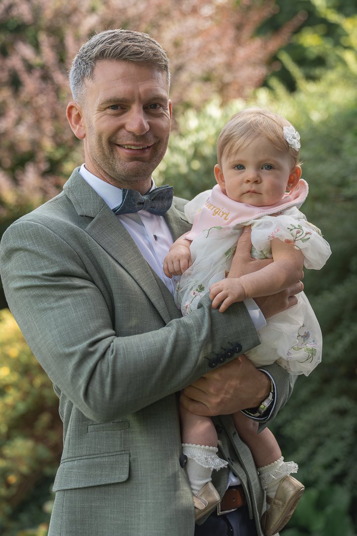 A man in a gray suit with a bow tie is holding a young girl in a white dress with floral embroidery. They are outdoors with trees in the background, and the man has a friendly smile on his face.