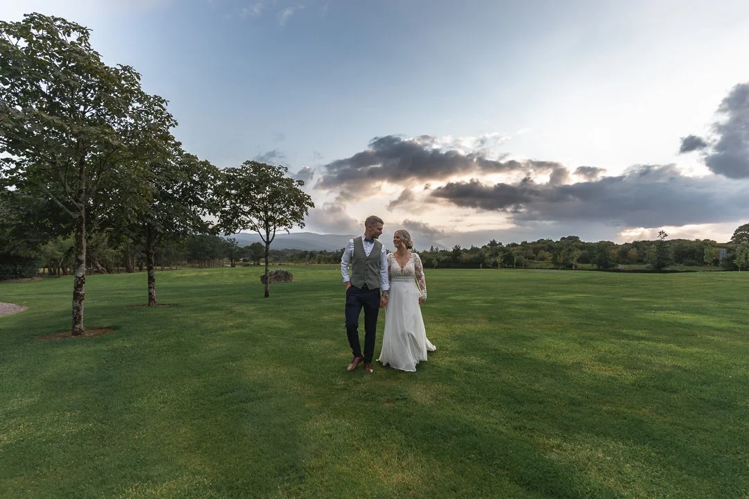 A bride and groom walking hand in hand on a lush green field during sunset, with trees and distant mountains in the background.