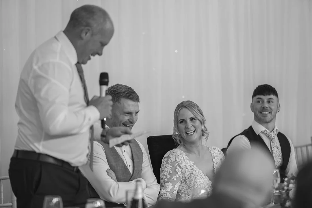 A wedding reception with four people seated at a head table, one man giving a speech, and the others laughing and smiling.