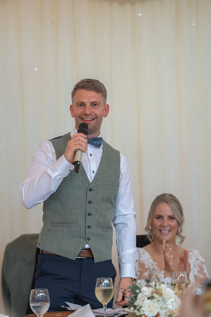 A man in a gray vest, white shirt, and bow tie is holding a microphone and smiling at a wedding reception. A woman in a white wedding dress is sitting beside him, clapping and smiling, with a floral centerpiece and glasses of white wine on the table.