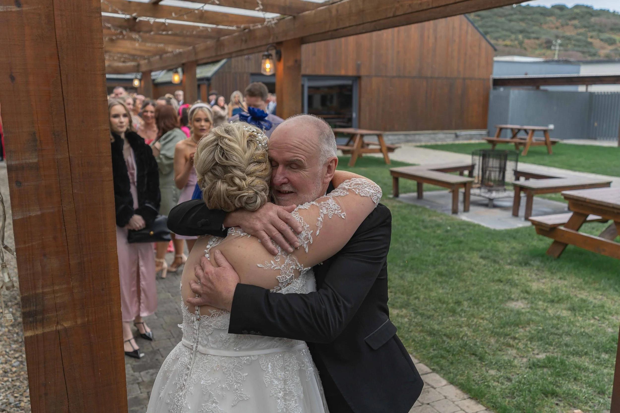 An elderly man embracing a woman in a wedding dress at an outdoor wedding reception, with guests watching in the background.