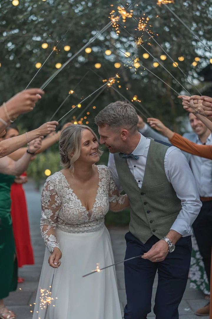 Bride and groom smiling and leaning into each other during a sparkler send-off at their wedding, surrounded by friends holding sparklers.