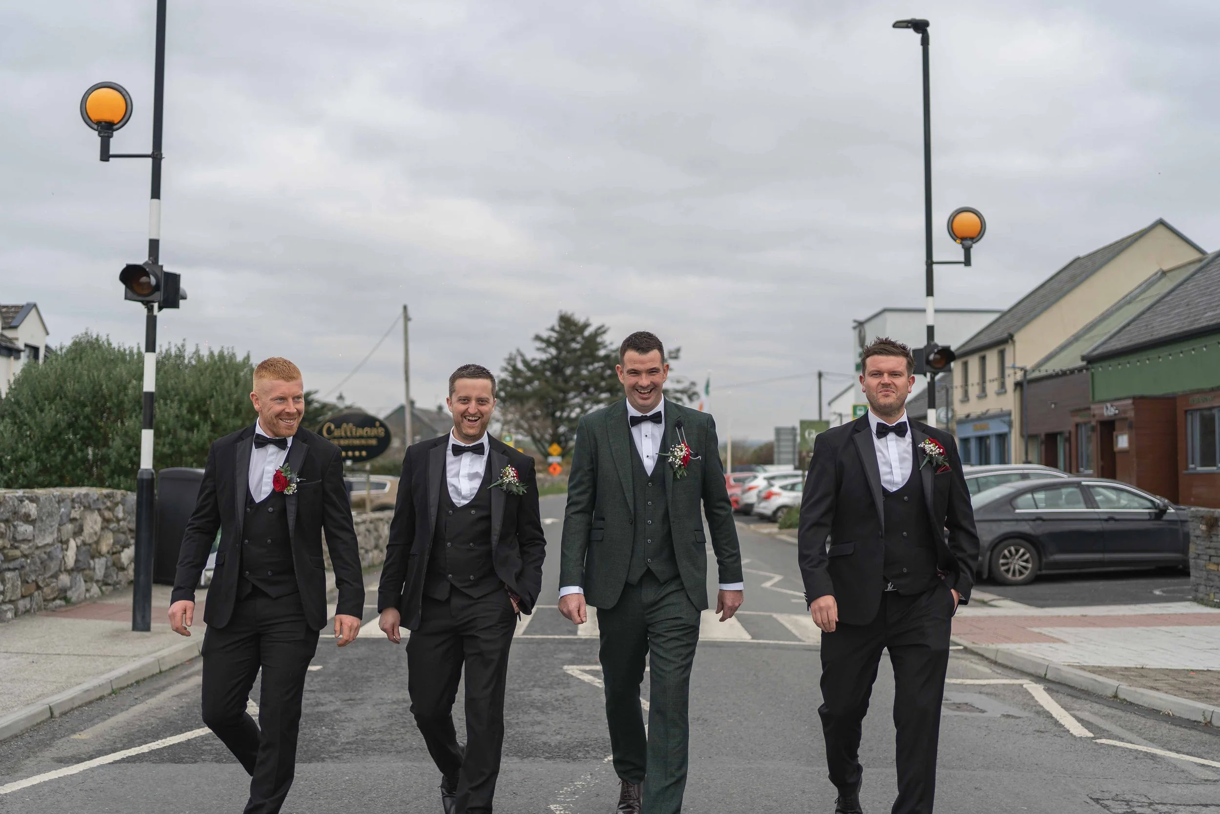 Four men in tuxedos walking on a street, smiling, with gray cloudy sky above, storefronts, parked cars, and streetlights visible in the background.