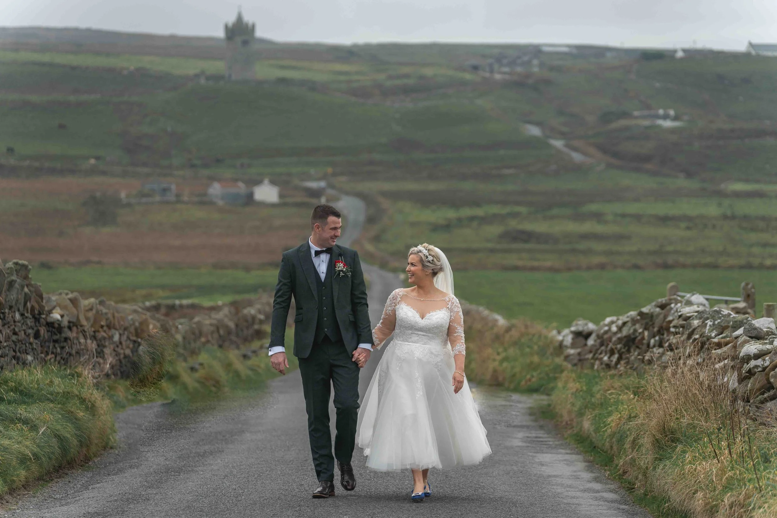 A bride and groom walking hand in hand on a rural road with stone walls, green fields, and rolling hills in the background, on a cloudy day.