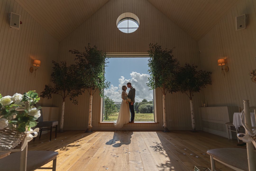 A couple in wedding attire standing on a platform inside a bright, wooden-walled chapel with large open window, trees, and blue sky with clouds outside, during daytime.