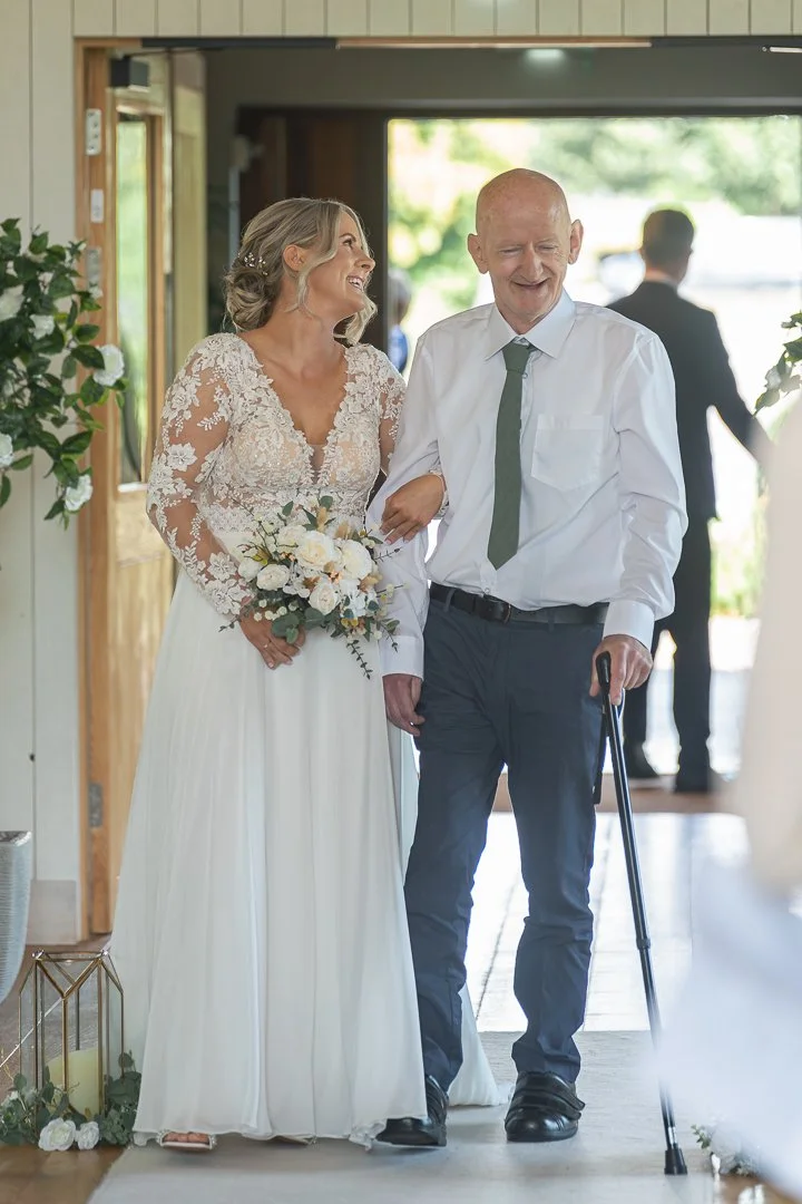 A bride in a lace wedding dress holding a bouquet, walking with an elderly man with a cane at a wedding ceremony.
