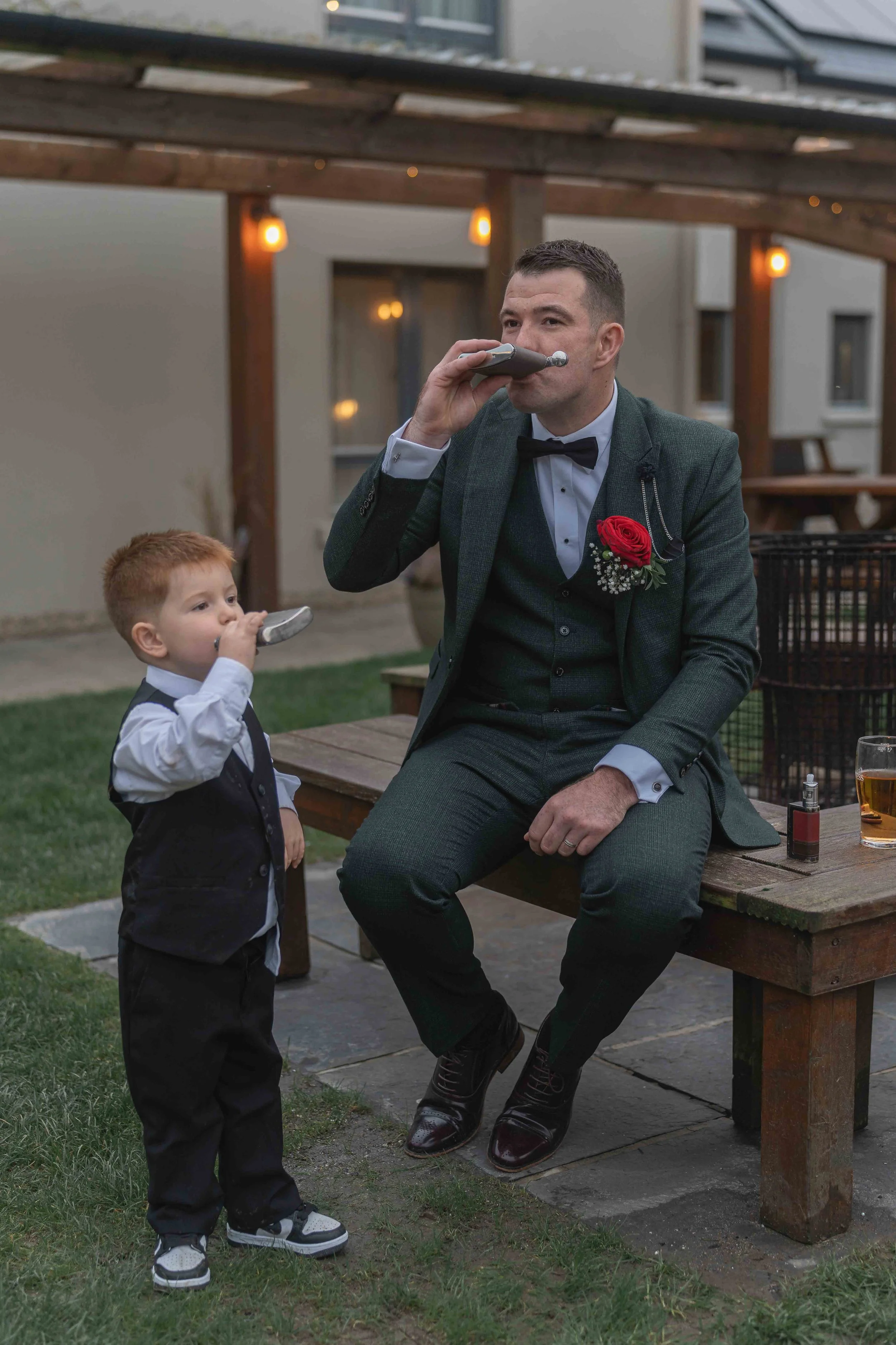 A man in a gray suit and black bow tie sitting on a wooden bench, and a young boy in a black vest, white shirt, and black pants playing a harmonica, in an outdoor setting with string lights and a modern building in the background.