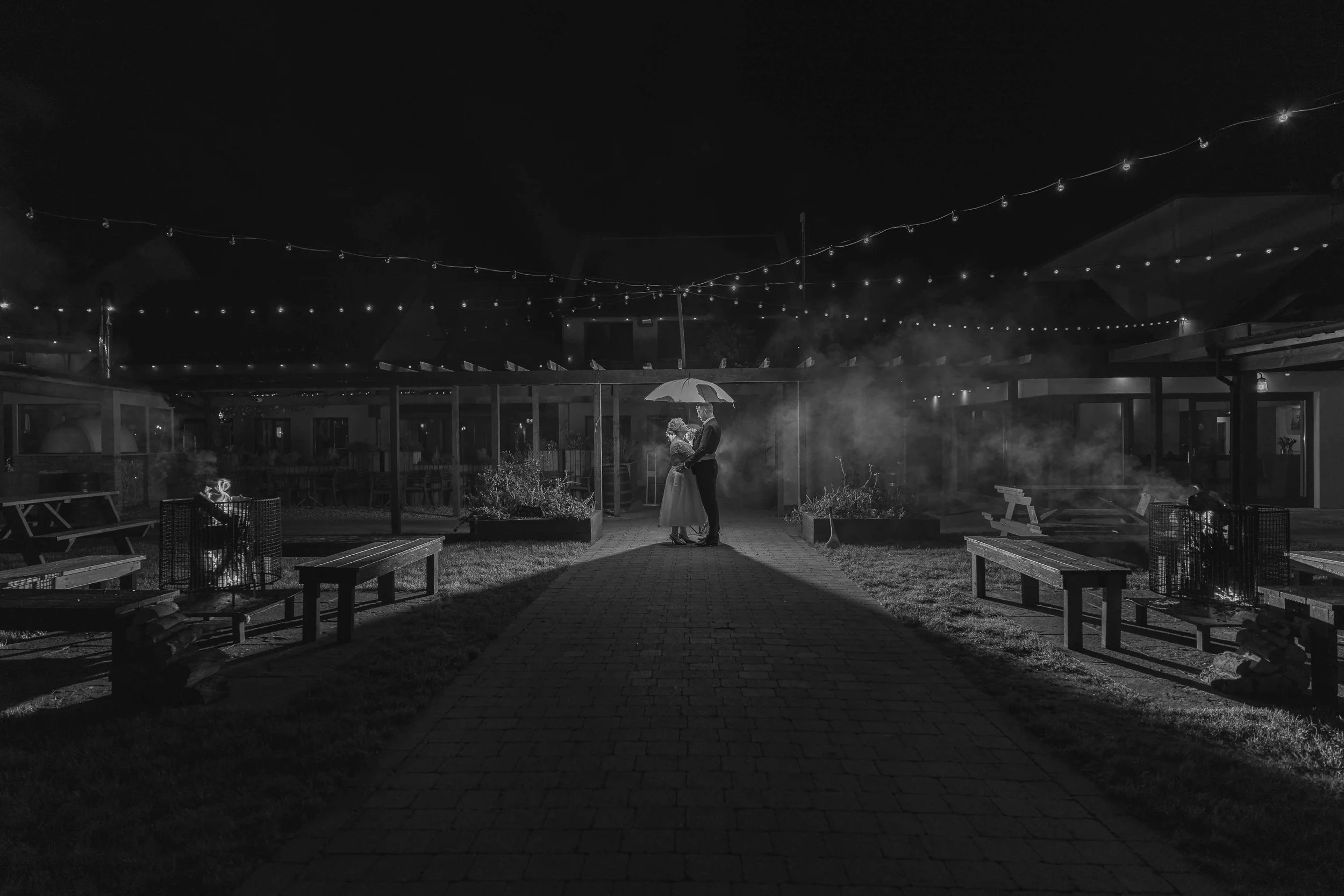 A couple standing under an umbrella, holding a bouquet, in an outdoor area with string lights overhead at night.