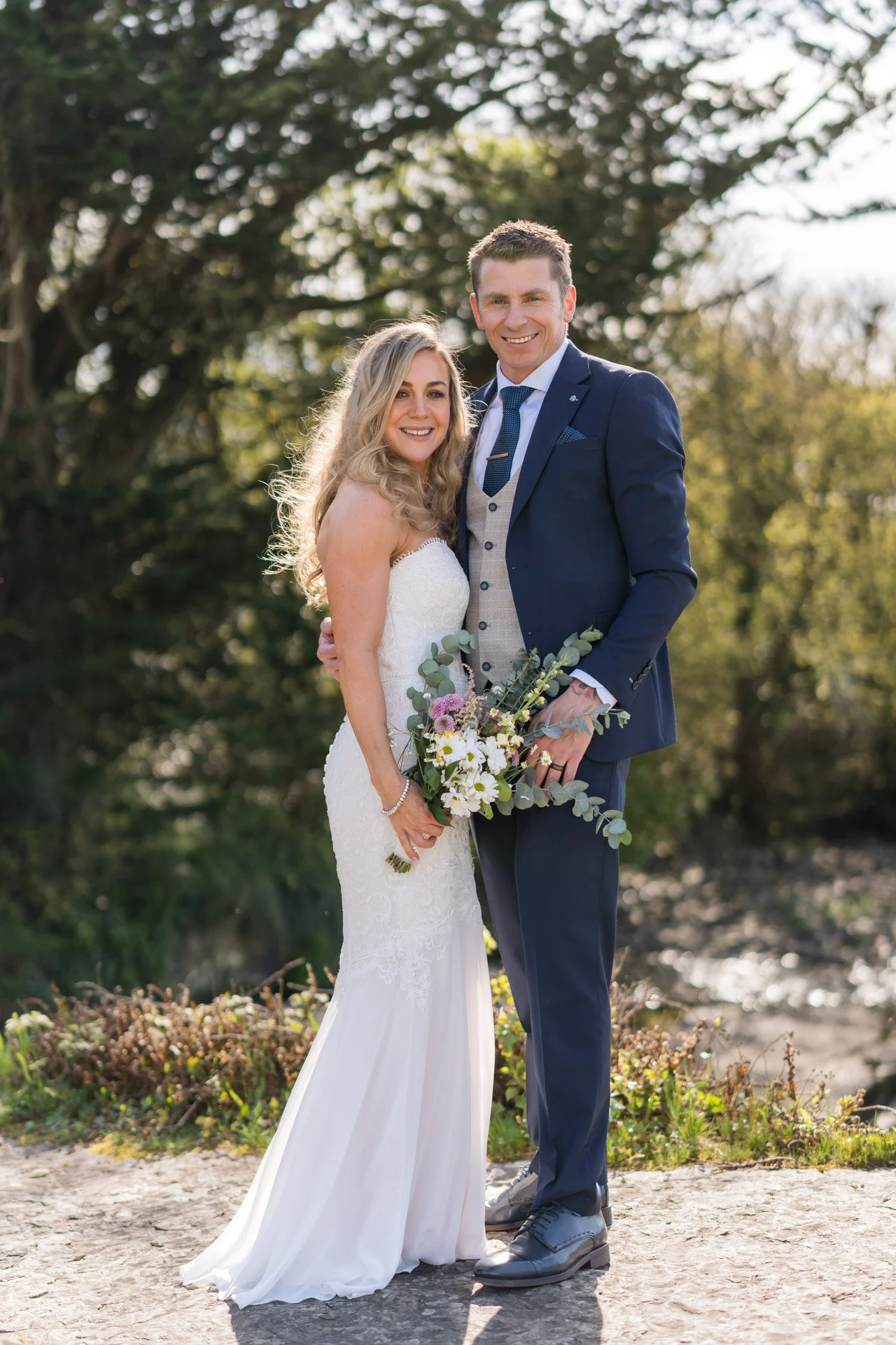 A newlywed couple stands outdoors on a dirt path, smiling. The bride wears a white lace wedding dress and holds a bouquet of flowers, while the groom wears a navy blue suit with a light gray vest and a blue tie. They are surrounded by greenery and trees, with sunlight filtering through the branches.