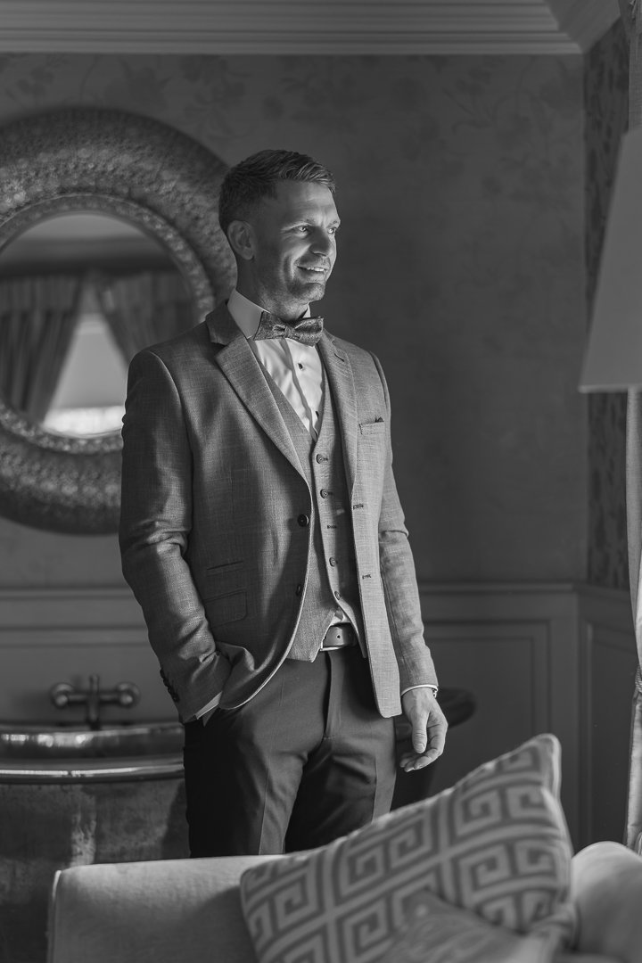 A man in formal attire, including a suit and bow tie, standing indoors near a mirror and furniture, smiling and looking to the side.