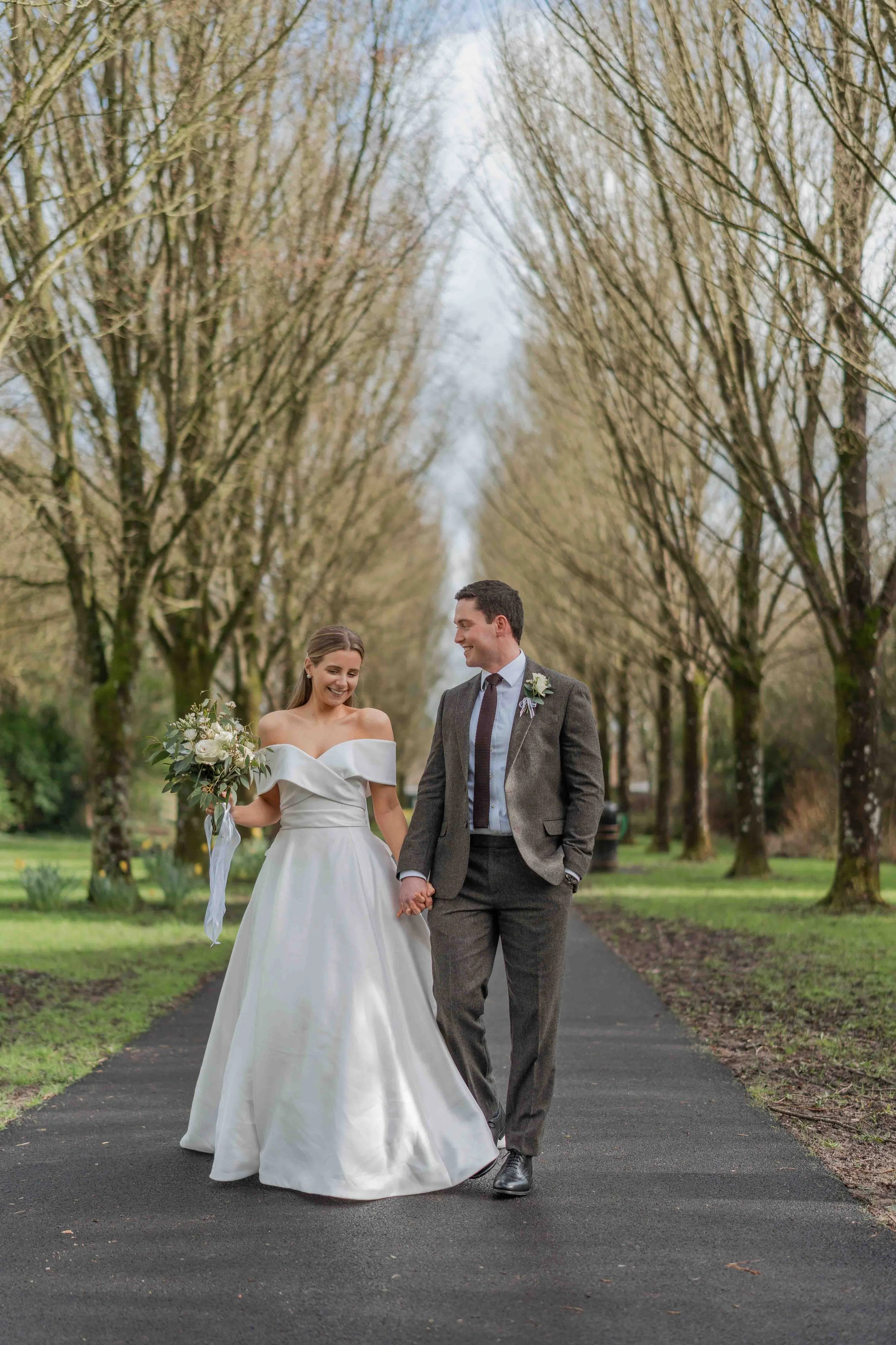 Bride and groom walking hand in hand on a tree-lined pathway, smiling and enjoying their wedding day outdoors.