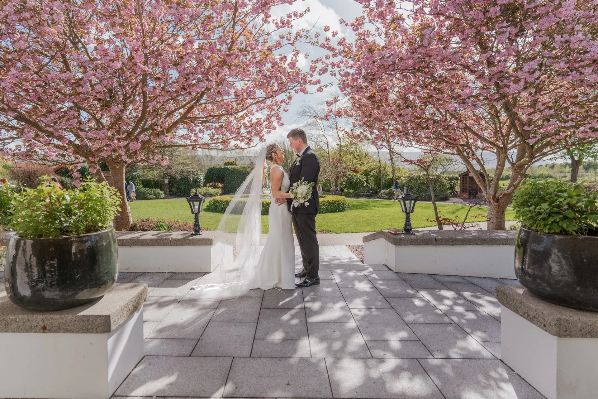 A bride and groom standing face to face under pink blossoming trees in a garden, holding hands, with the bride holding a bouquet of white flowers.