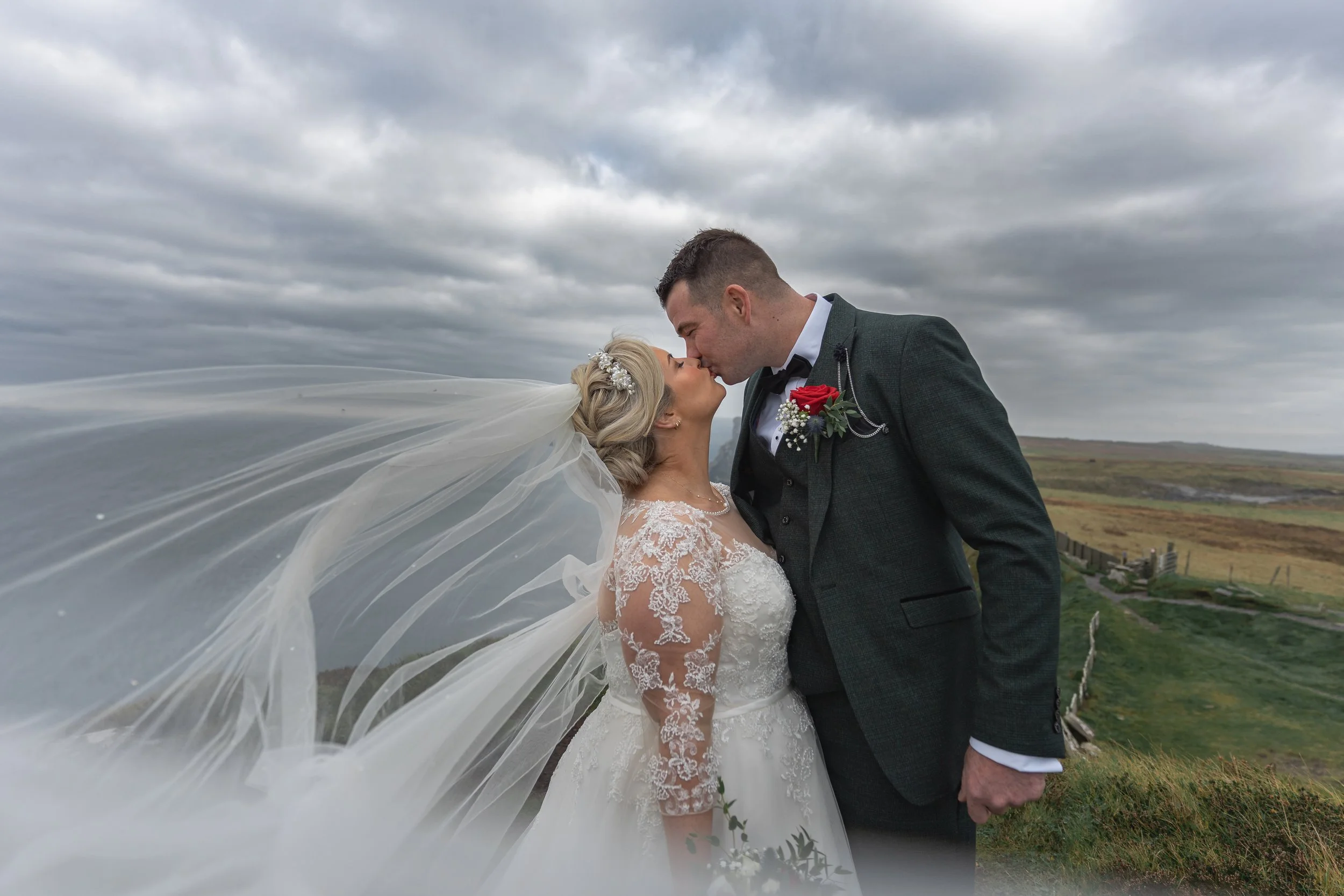 A bride and groom are kissing outdoors on a cloudy day, with the bride wearing a lace wedding dress and a flowing veil, and the groom in a dark suit with a red boutonniere, on a grassy hillside.