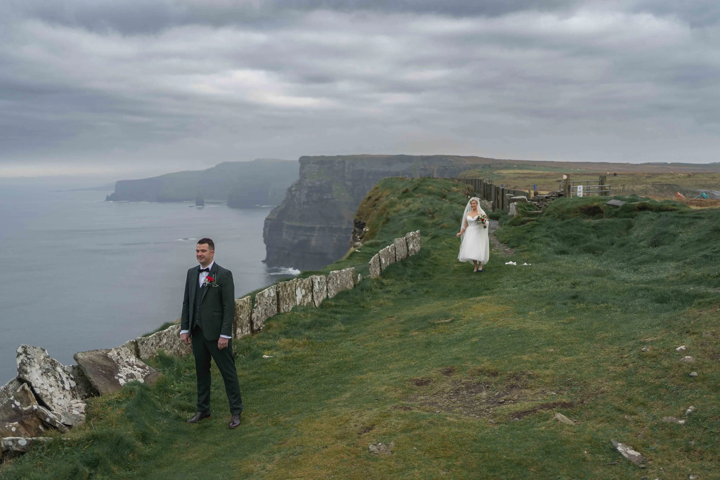 A bride and groom standing on a grassy cliffside with dramatic ocean and rocky cliffs in the background. The groom is dressed in a formal black suit with a red boutonniere, and the bride is in a white wedding dress holding a bouquet of flowers.