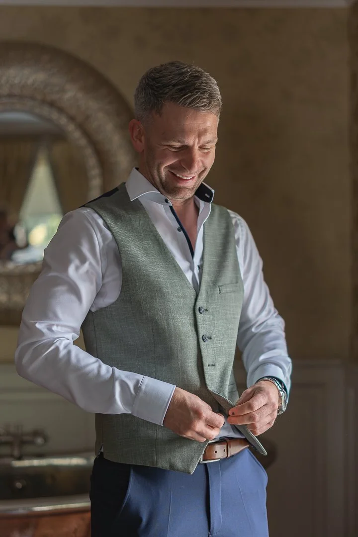 A man with short gray hair wearing a white dress shirt, gray vest, and blue pants, smiling and adjusting his vest in a warmly lit room with a decorative mirror behind him.