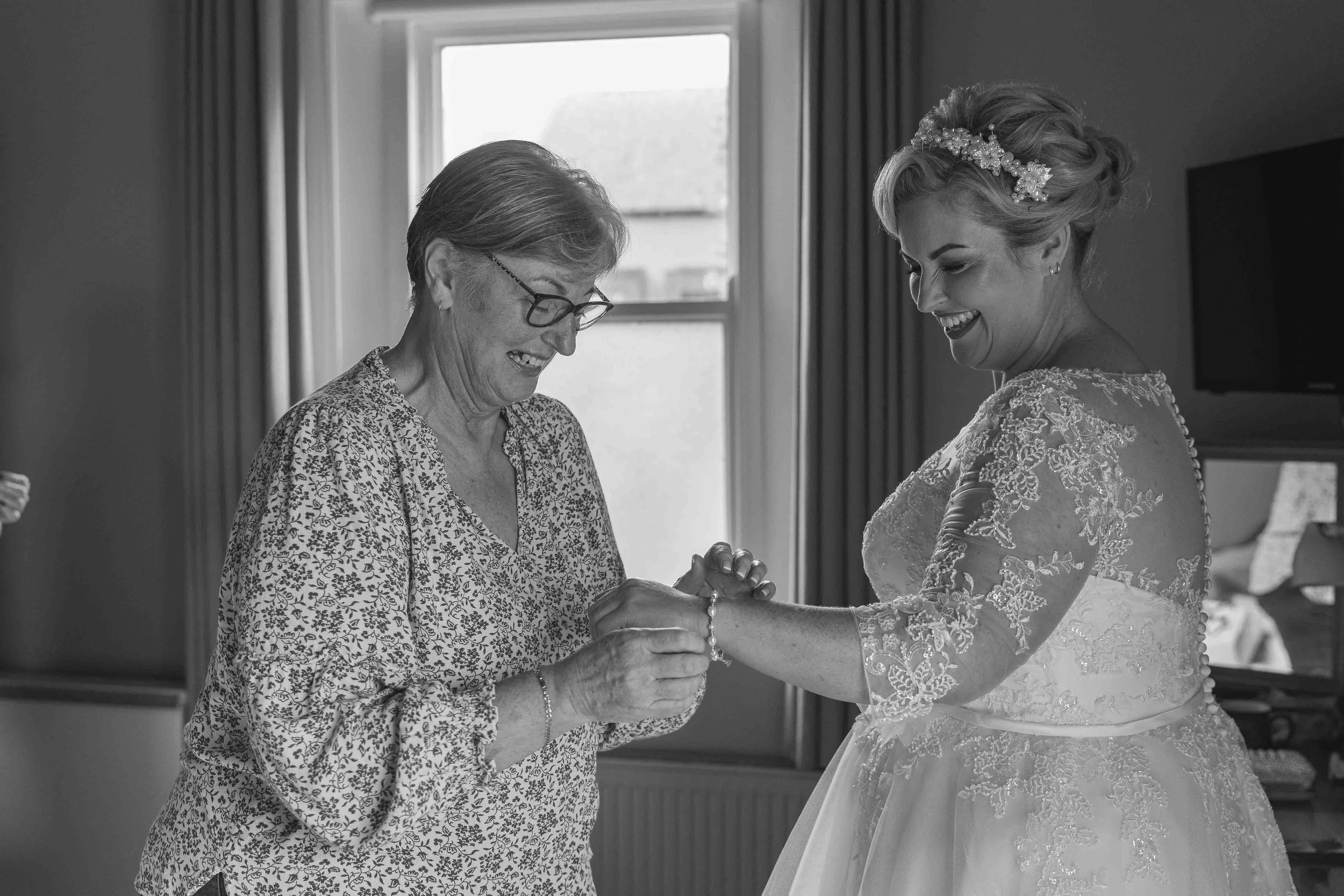 A bride with floral lace wedding dress and floral headband kisses her elderly mother on the hand inside a room with a window and curtains.