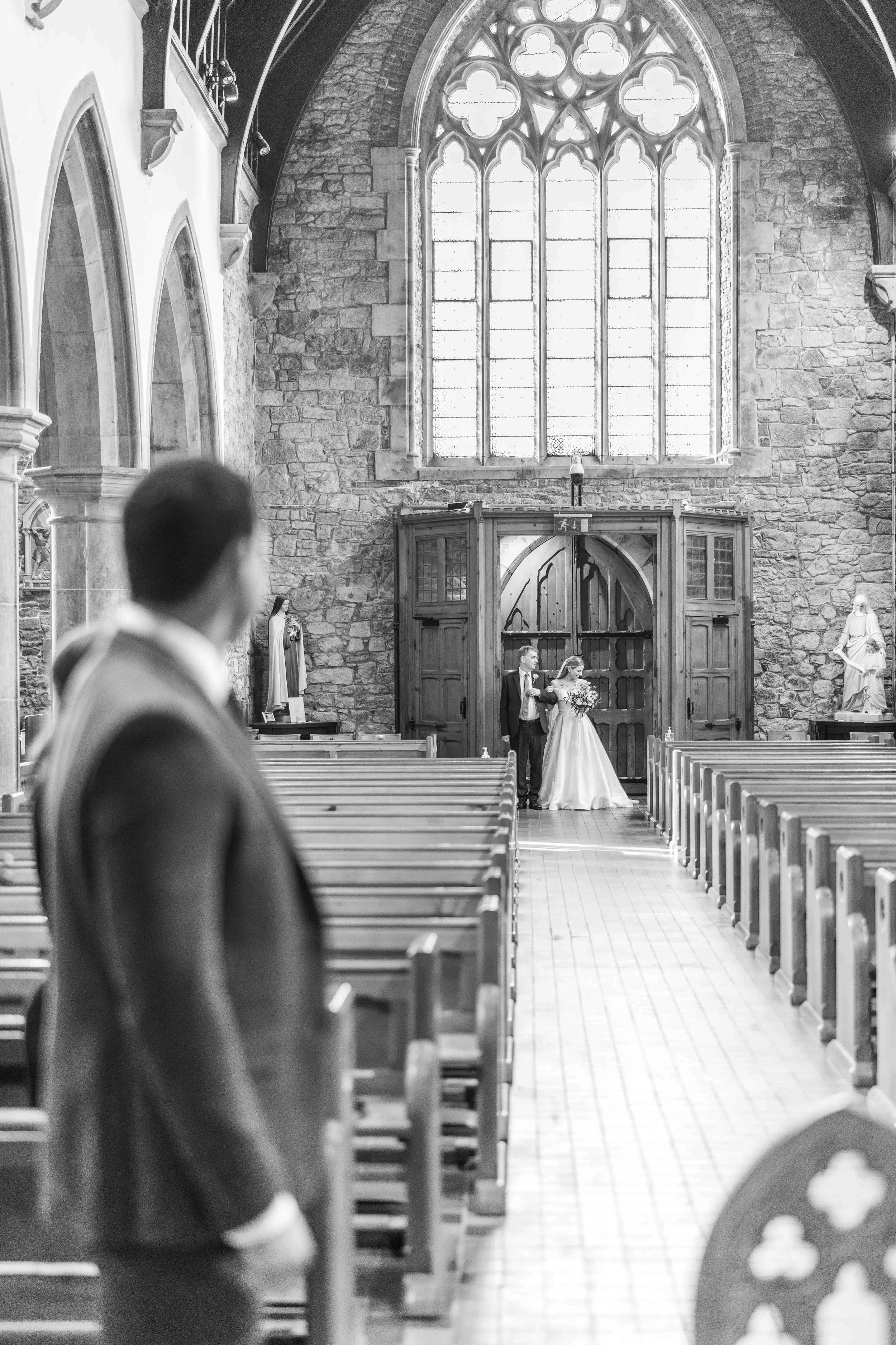 Black and white photo of a wedding ceremony in a church with a large stained-glass window at the front. A bride is walking down the aisle with a person, possibly a family member, while the groom waits at the altar. The groom is in focus in the foregr