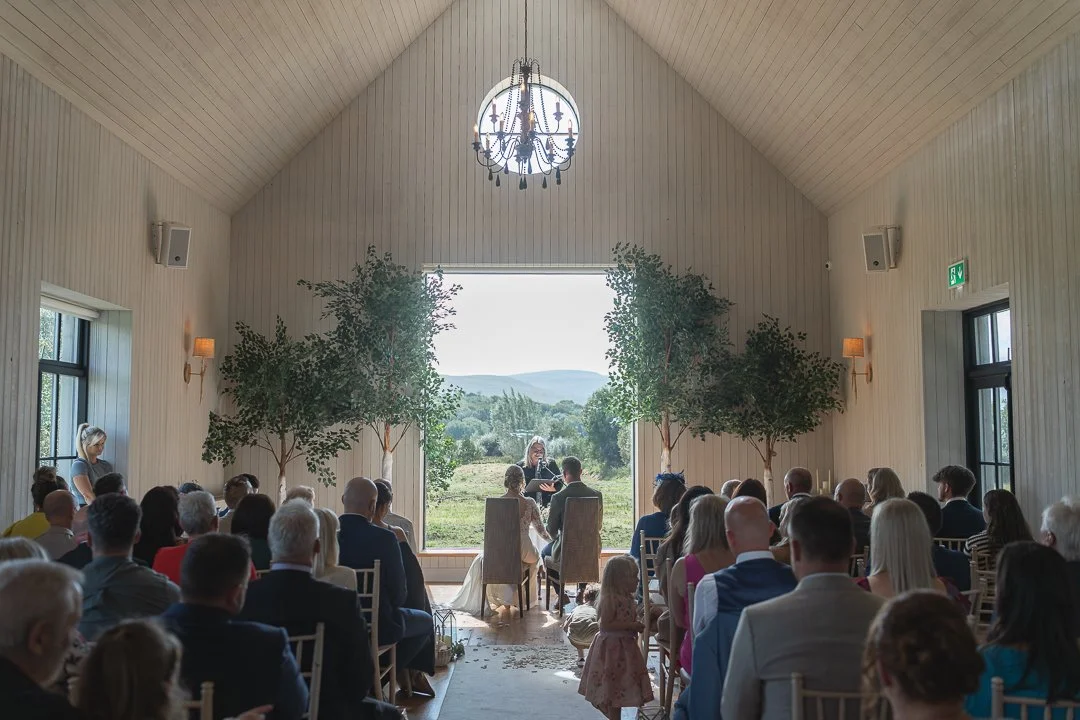 Wedding ceremony inside a white wooden chapel with large open doors showing a scenic outdoor view, surrounded by trees. Guests are seated facing the couple, who are sitting at the front with an officiant. A chandelier hangs from the vaulted ceiling, 