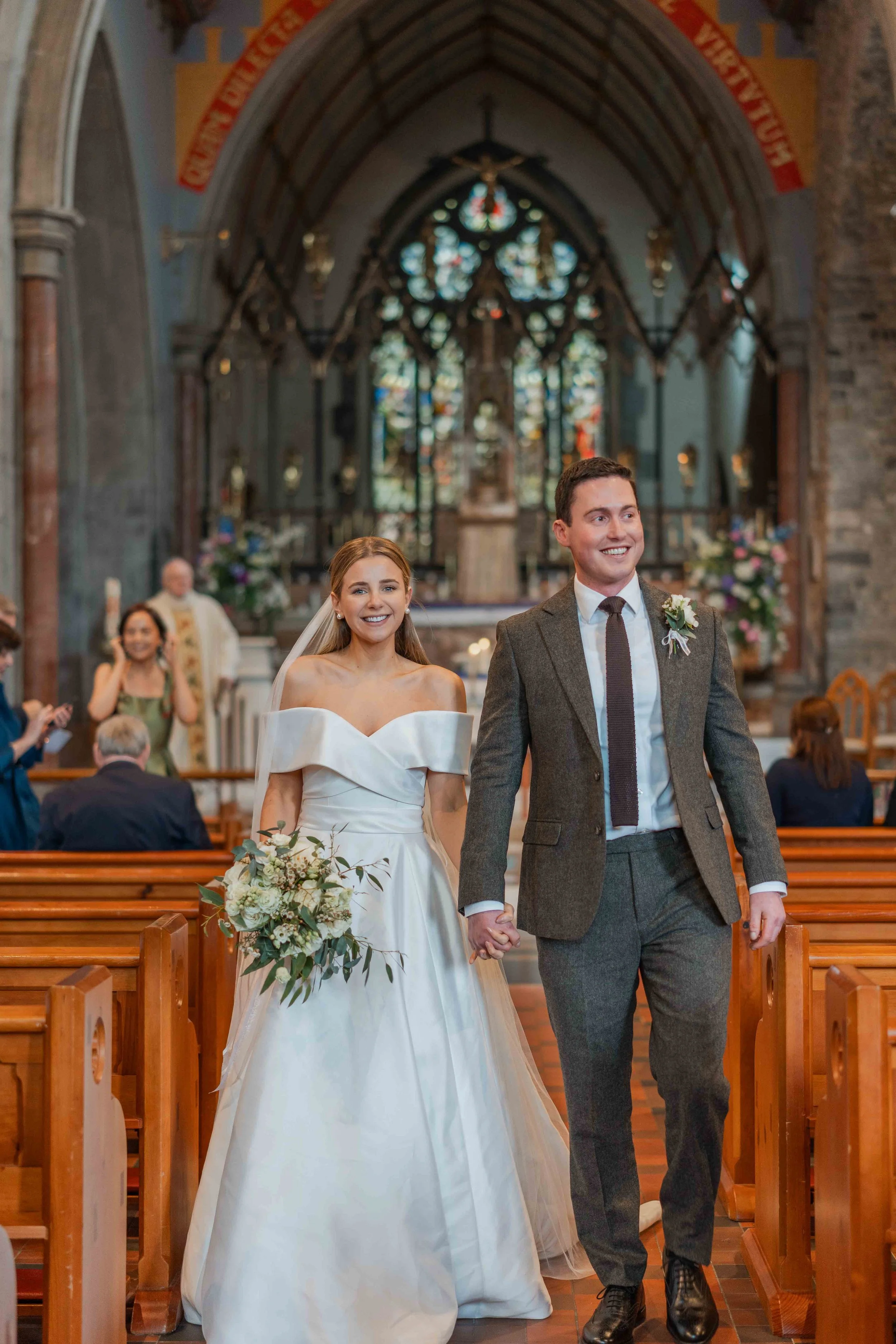 A newly married couple walking down the aisle in a church, holding hands and smiling, with guests seated on pews and colorful floral arrangements in the background.
