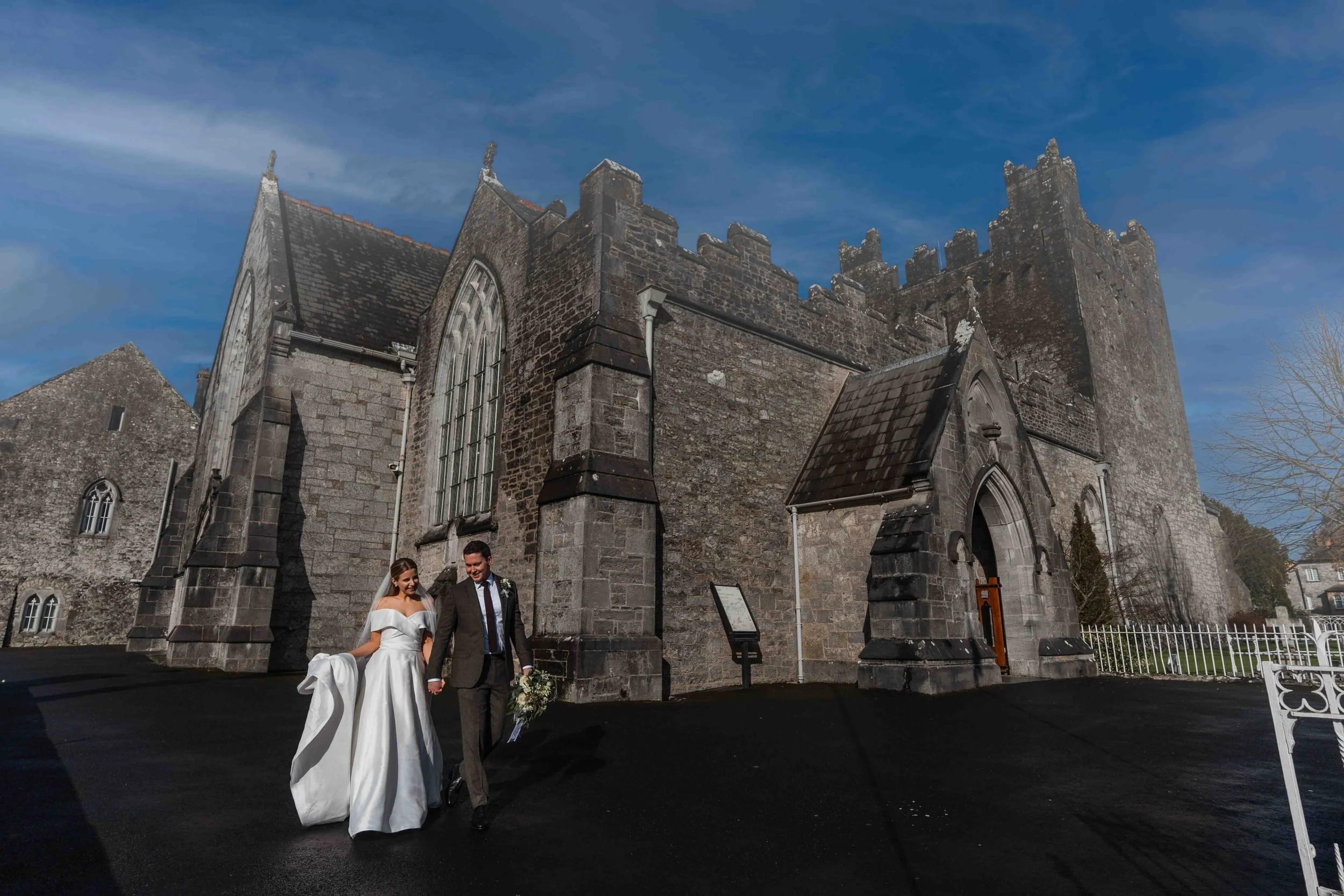 A bride and groom walking hand in hand outside a historic stone church on their wedding day, with blue sky and clouds in the background.
