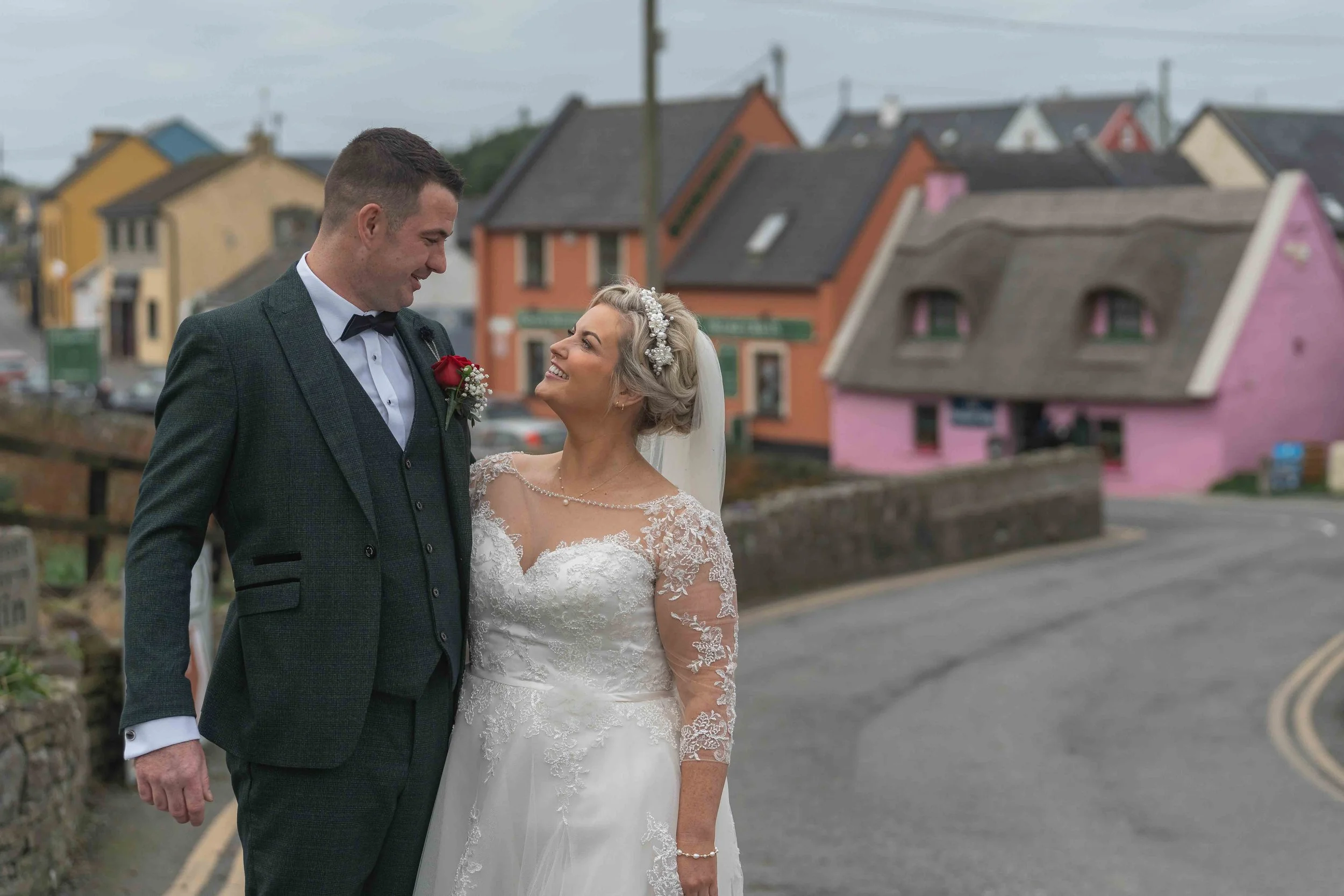 A bride and groom in wedding attire standing close together on a street, smiling at each other with colorful houses in the background.