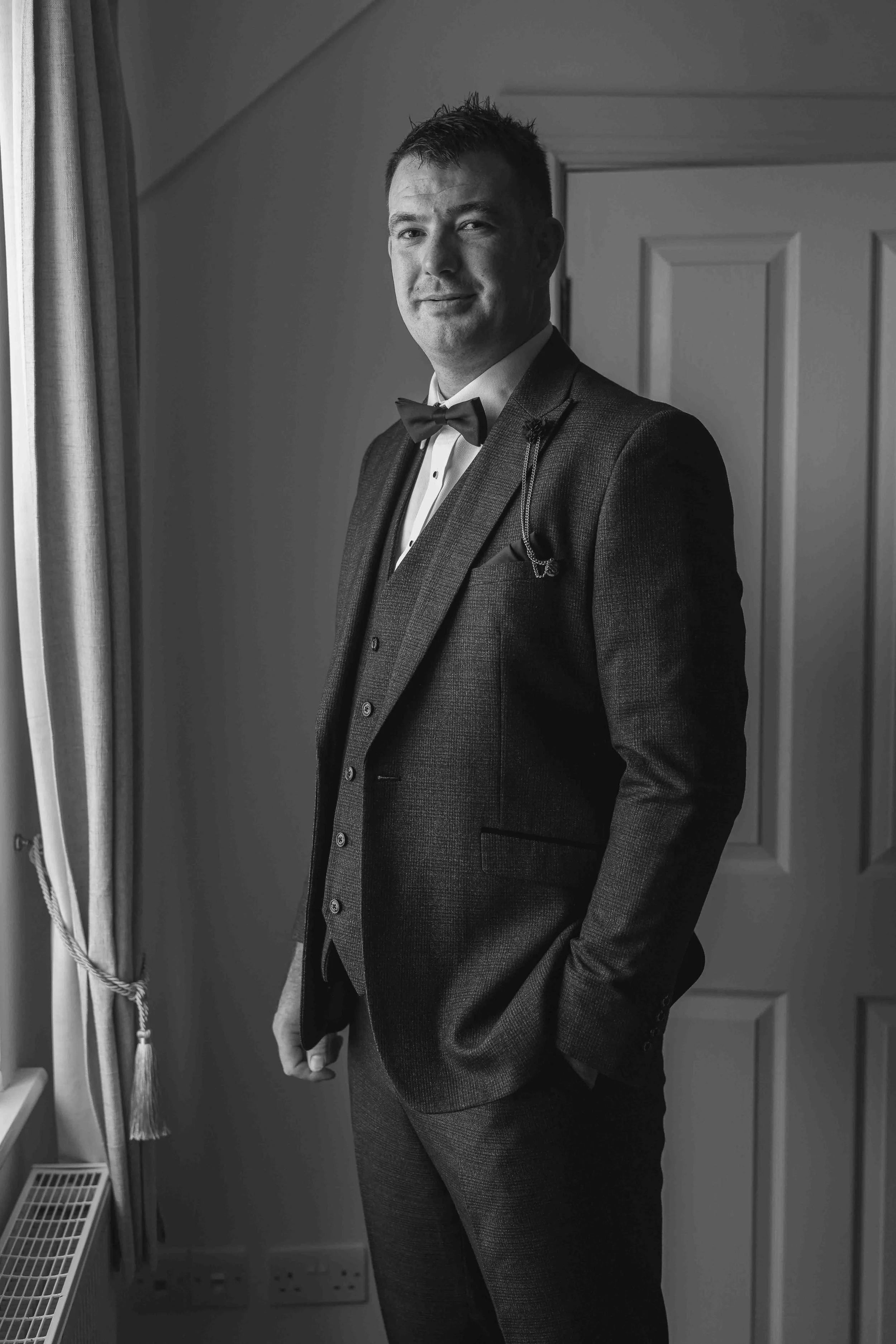 A man in a tuxedo standing indoors next to a window with curtains, smiling slightly, in front of a closed door.