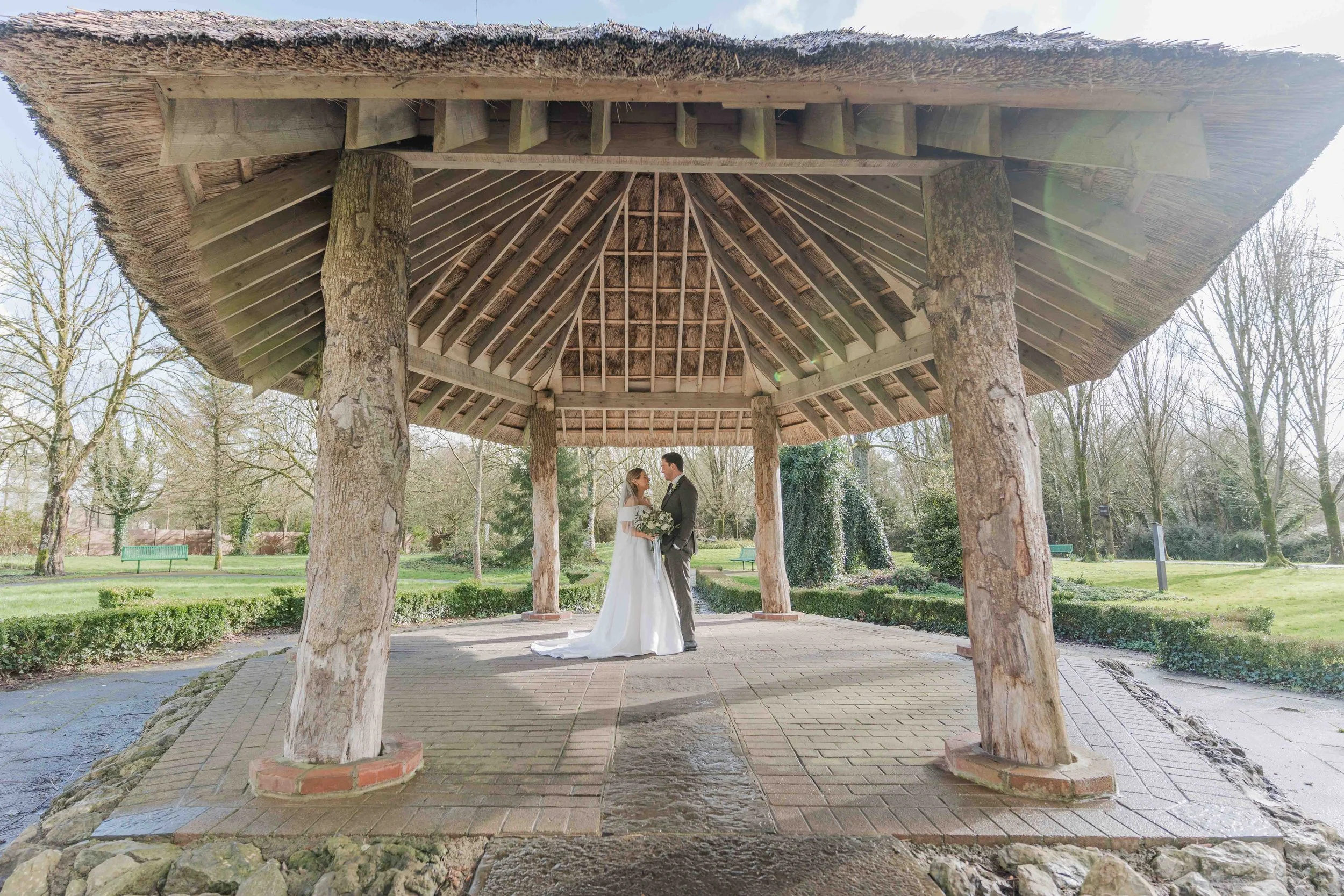 A bride and groom standing under a wooden gazebo in a park, holding hands and looking at each other.