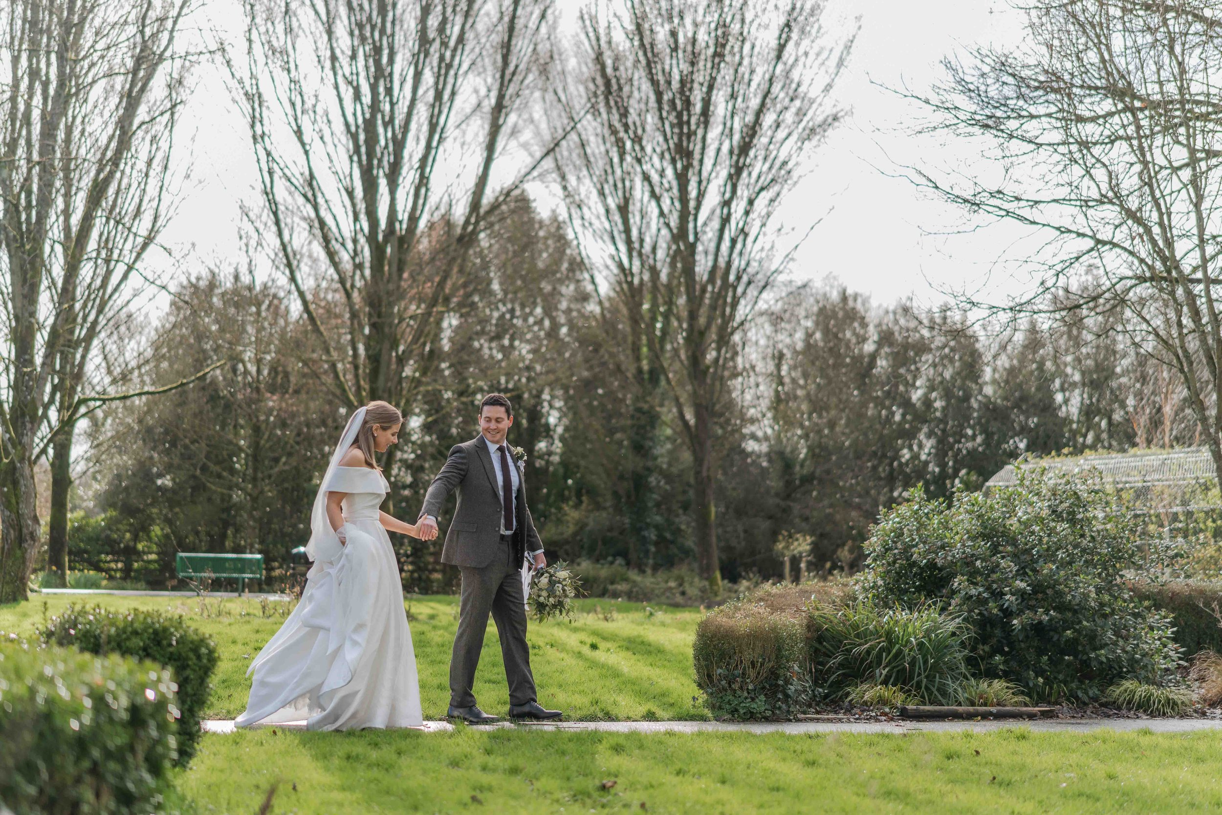 A bride and groom walking hand in hand outdoors in a green park during daytime.