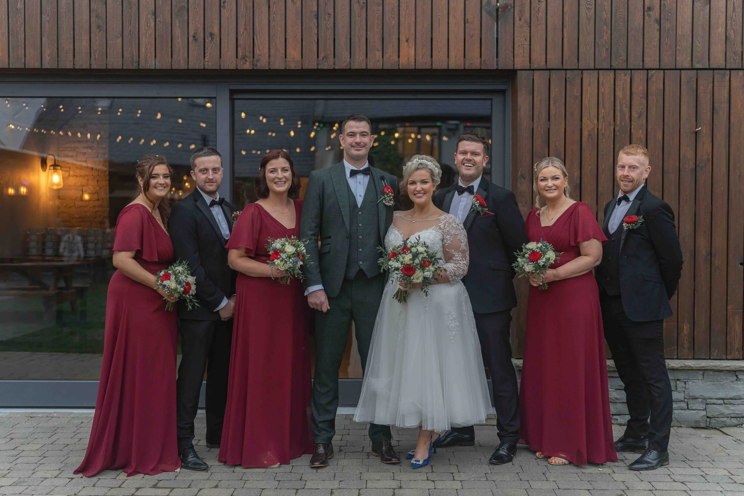 A wedding party of nine people standing outside in front of a wooden building with a glass door. The bride and groom are in the center, the bride wearing a white wedding dress and holding a bouquet, the groom in a gray suit with a bow tie. Four women