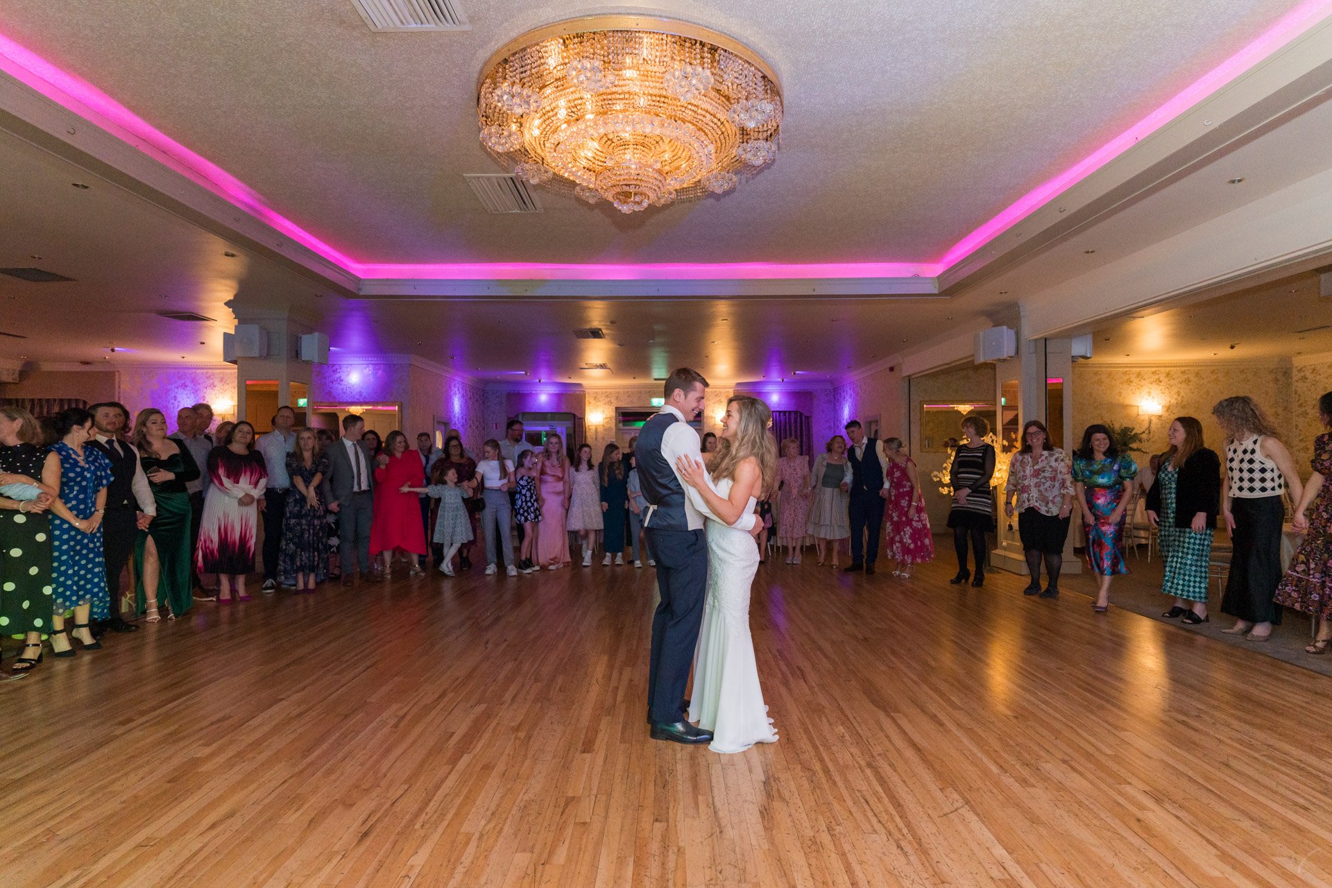 A bride and groom dancing in the center of a wedding reception hall, surrounded by guests watching.