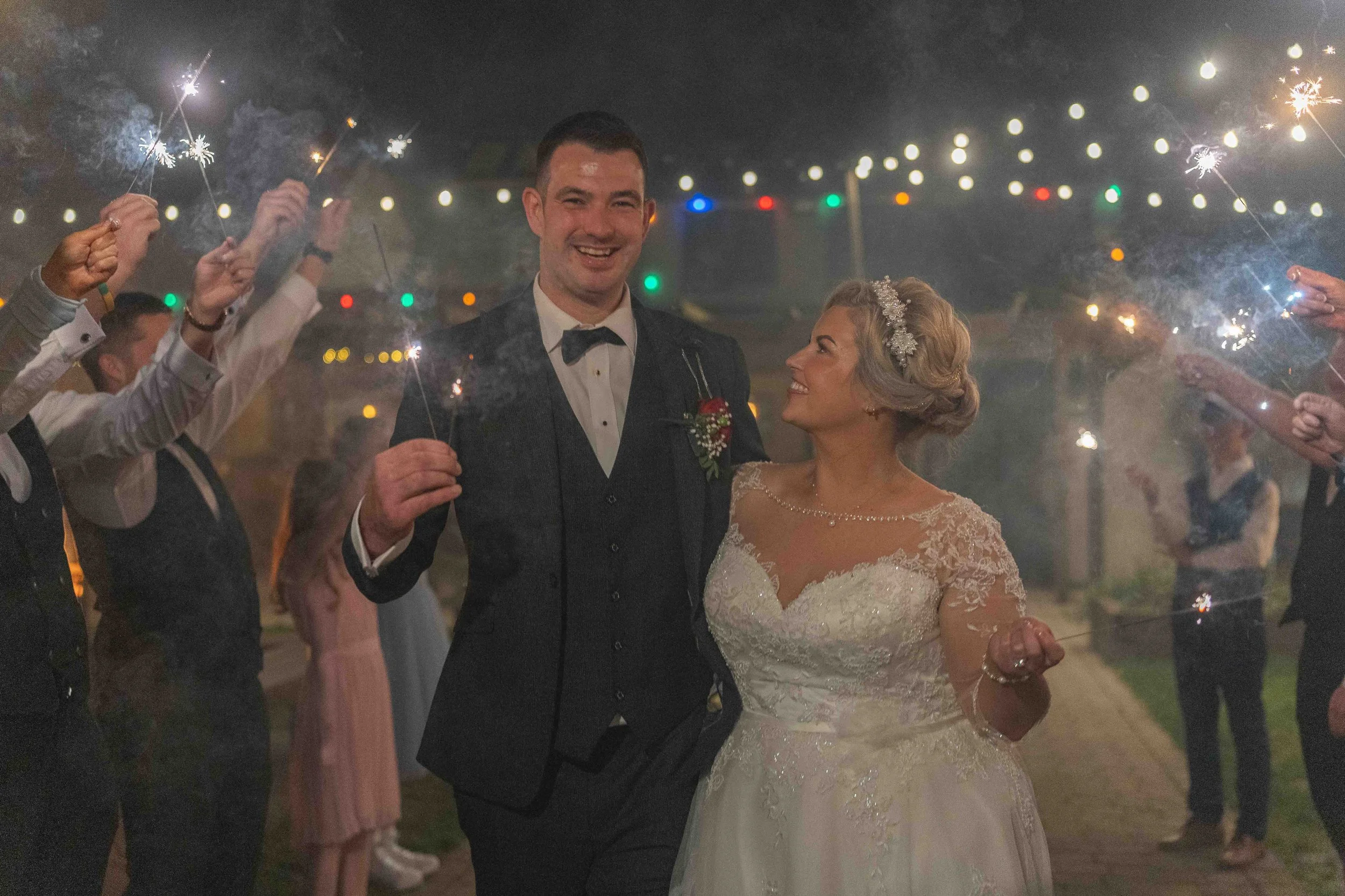 A bride and groom at their wedding reception, celebrating with sparklers held by guests under string lights at night.