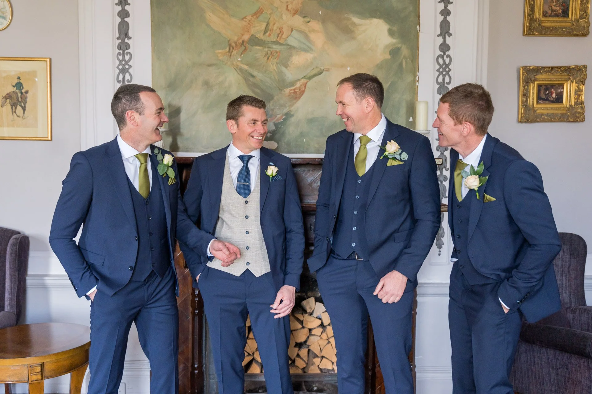 Five men in suits standing together indoors, smiling and talking, with a fireplace and decorative artwork behind them.