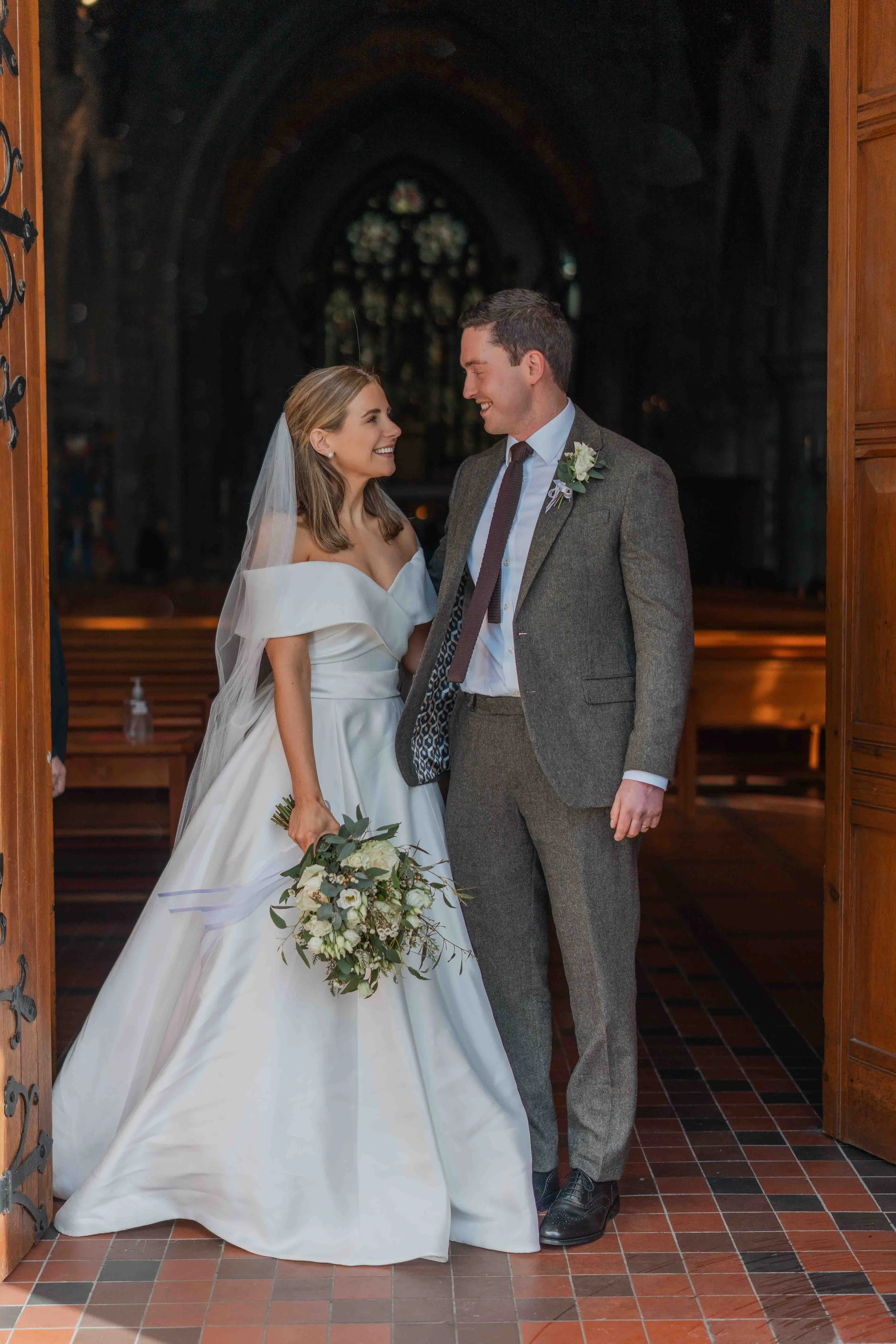 A bride and groom standing inside a church, smiling and looking at each other, with the bride holding a bouquet of flowers.