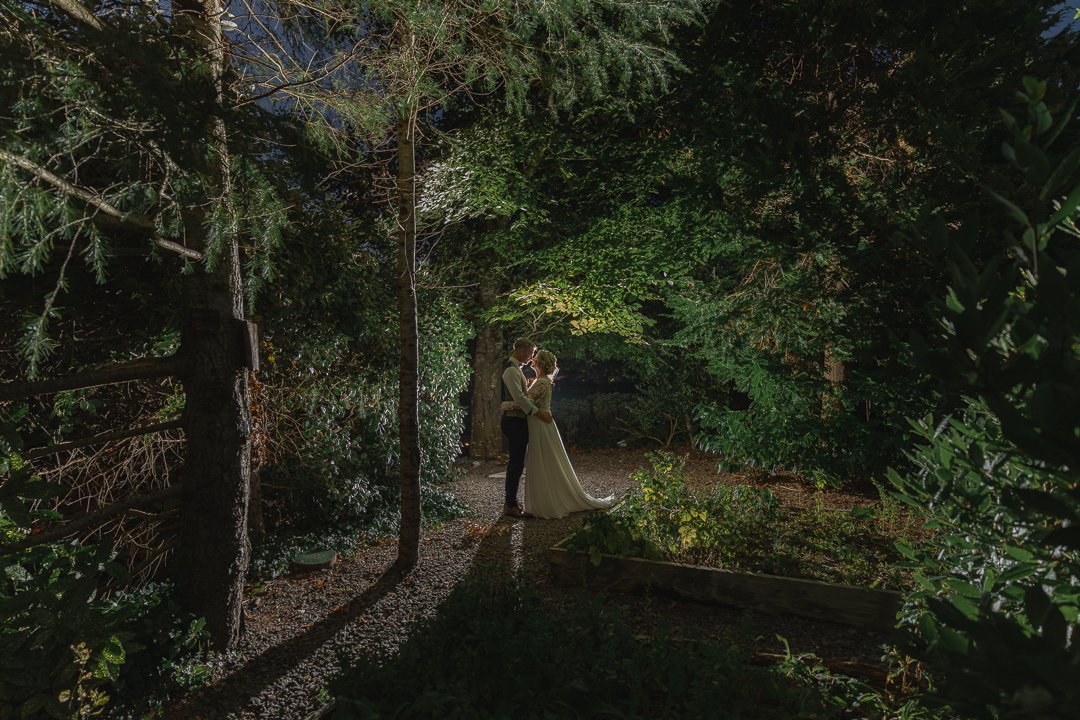 A couple sharing a kiss in a lush, green garden at night, illuminated by soft lighting.
