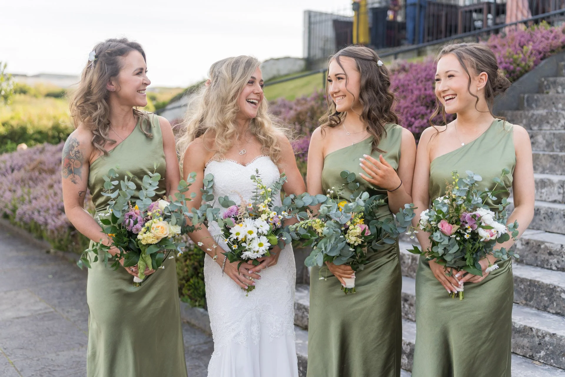 A bride and four bridesmaids standing together outdoors, all smiling and holding bouquets of flowers, with stairs and purple flowers in the background.