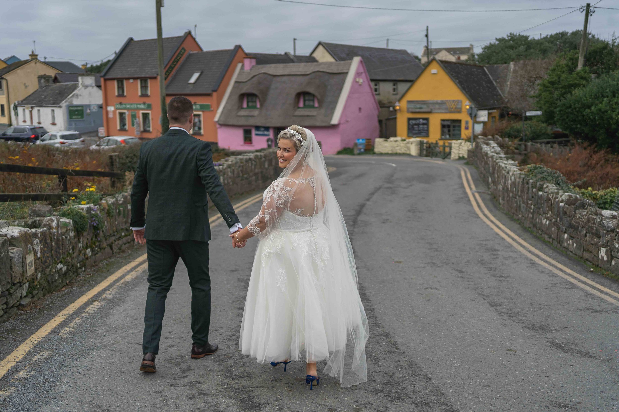 A bride and groom holding hands, walking down a street in a small town with colorful houses, the bride wearing a white wedding gown and veil, looking back and smiling.